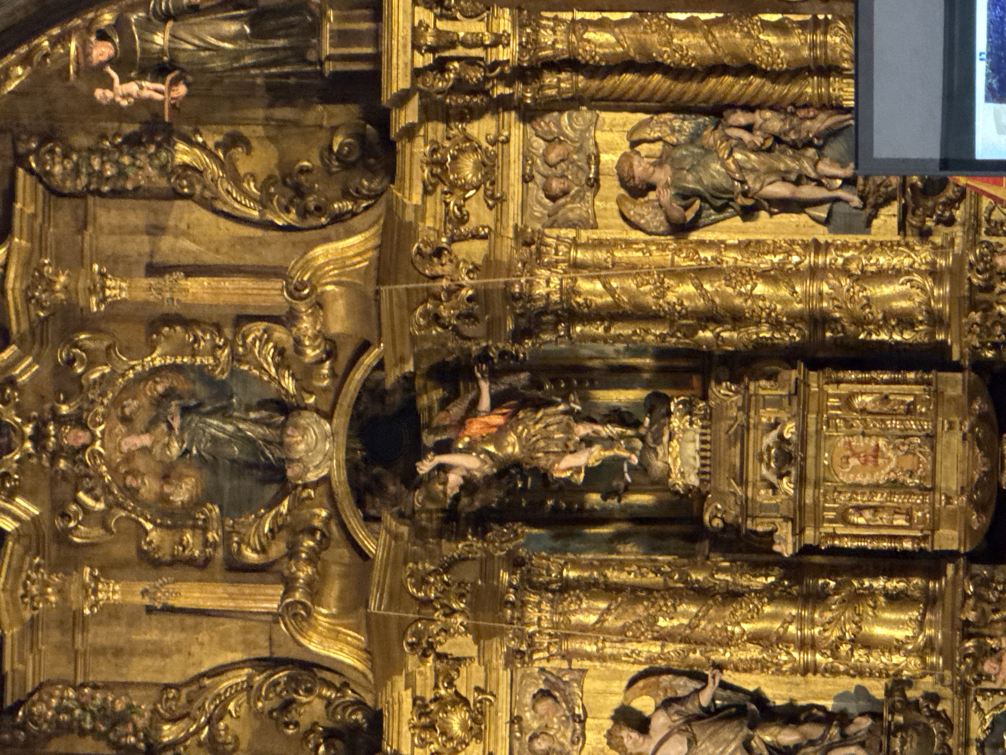 The image shows an ornate golden altar within a church in Palma de Mallorca, Spain. The altar is elaborately decorated with carvings and gilded sculptures. In the central upper section, a circular depiction features a figure of Mary holding a child, surrounded by smaller figures. To the right, a sculpture of a robed monk holds a small, nude child. The main feature of the altar is a large sculpture of an angel in dynamic pose, with outstretched wings and a flowing garment, holding a sword. To the left and right of the central angel, there are further sculptures of figures, some winged and robed, set within gilded architectural niches. The entire structure is characterized by intricate scrollwork, columns, and decorative elements, bathed in warm, artificial light that accentuates the golden sheen of the artwork.