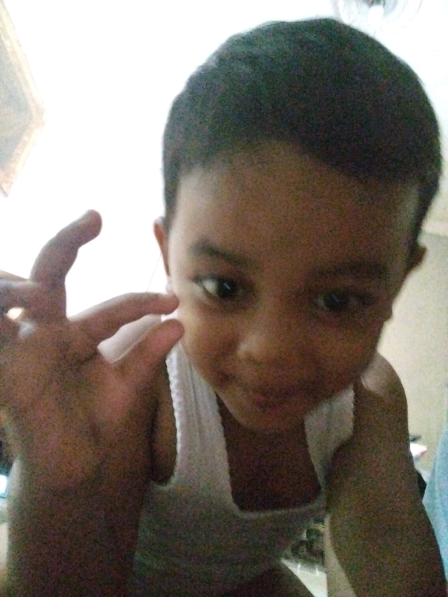 In Pepelegi, Indonesia, a close-up, low-angle selfie shows a young child, likely male, with dark hair and brown eyes, looking directly at the camera with a playful expression. The child is wearing a white ribbed tank top. Their left hand is raised, with fingers extended and forming an 'OK' gesture. The background is indistinct and brightly lit, suggesting an indoor setting. The image has a slightly grainy quality, typical of older digital photography.