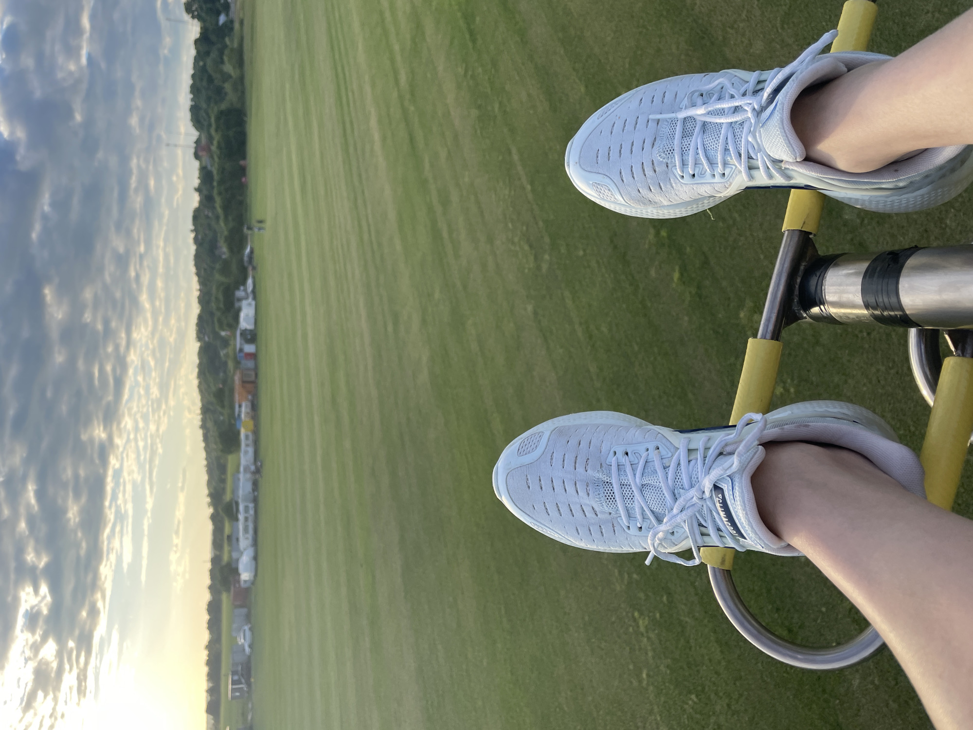 The image shows a first-person perspective of someone's feet wearing white athletic shoes, positioned on a metal and yellow-bar apparatus. The feet are against a backdrop of a vast, well-maintained green field with striped patterns from mowing. In the distance, a row of mobile homes or trailers is visible, along with some trees and structures. The sky above is partly cloudy with sunlight breaking through, indicating it might be late afternoon or early morning. The location is identified as Турово, Russia.