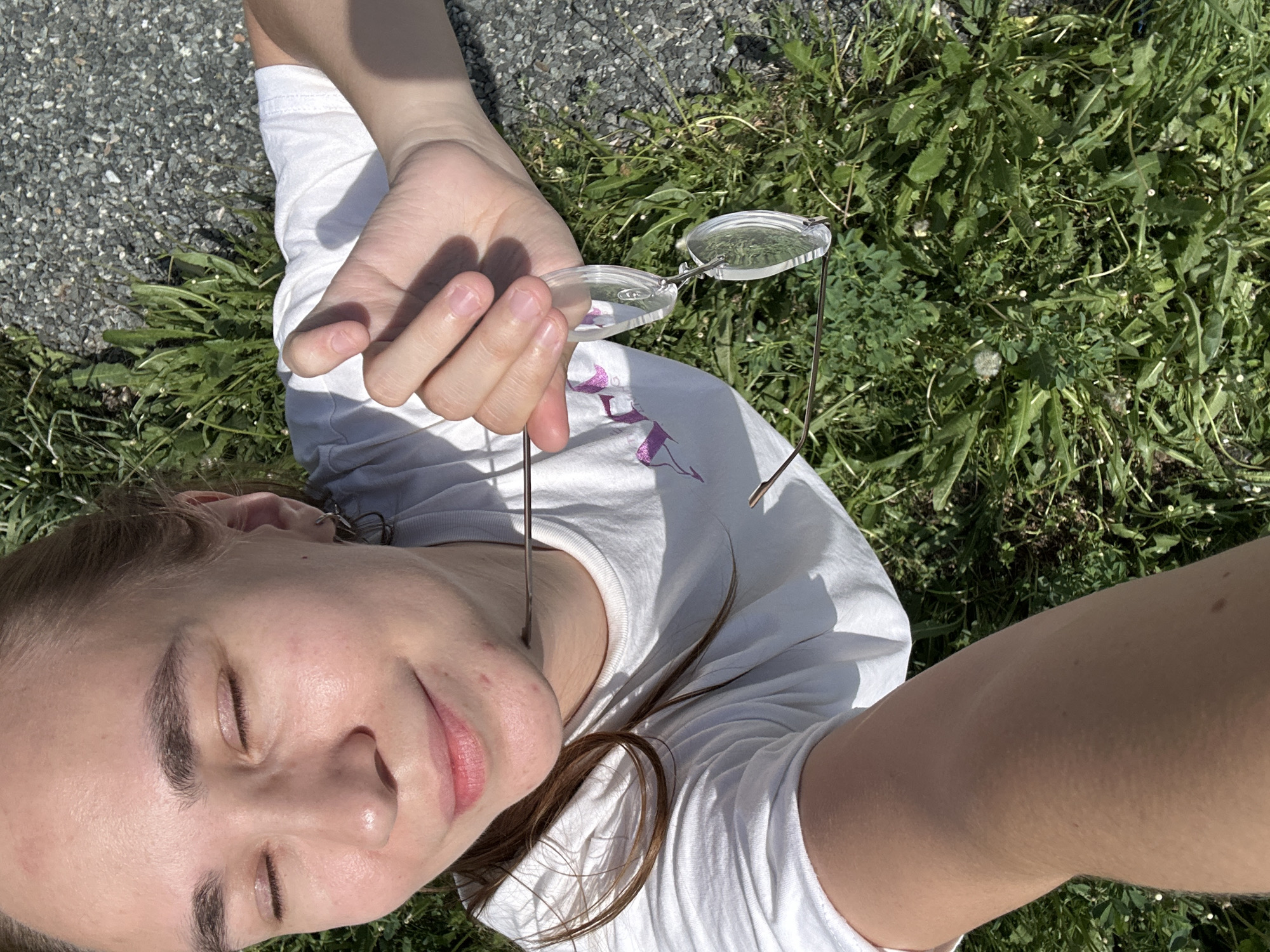 A fair-skinned young adult with closed eyes is lying on the ground, which consists of asphalt and grass, holding a pair of frameless eyeglasses with metal arms. The person is wearing a white t-shirt with a faint pink or purple design on the front. The location is unknown and cannot be identified.