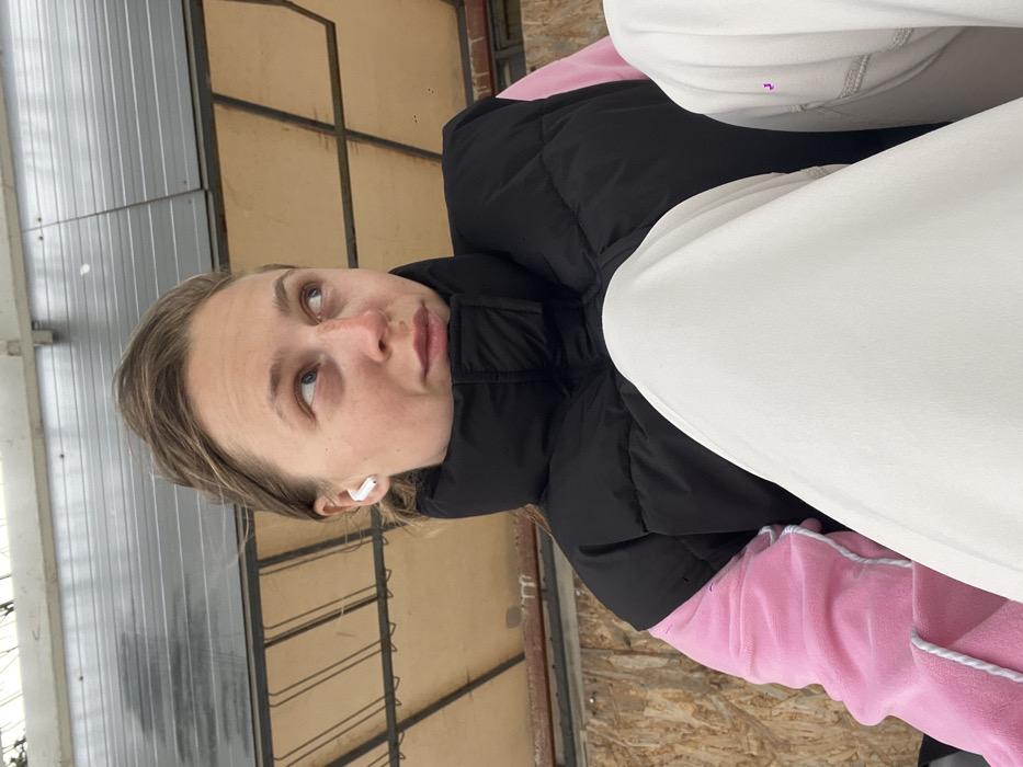 A young adult female is depicted in a medium shot, looking upwards and to the viewer's right with a neutral expression. Her light brown hair is pulled back. She exhibits freckles on her nose and cheeks, and a white wireless earbud is present in her left ear. She is wearing a black high-collar puffer vest over a pink long-sleeved garment. White or light-colored trousers are partially visible in the lower foreground. The background consists of a grey corrugated metal ceiling or overhang above a section of light-colored wooden panels arranged in a grid pattern. The specific location within Budva, Montenegro, is not identifiable from the visible architectural elements.