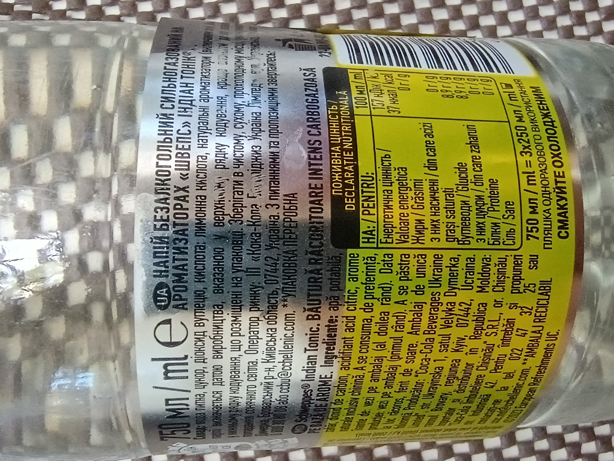 The image shows a close-up of a glass bottle of "Schweppes Indian Tonic" beverage. The bottle is mostly transparent, revealing the clear liquid inside. The label wraps around the bottle, displaying a significant amount of text in Ukrainian and Romanian, along with a barcode and nutritional information. The label is a mix of silver, yellow, and black, with some text highlighted in bright colors.
The text on the label indicates that the beverage is "non-alcoholic, carbonated, with natural flavourings" and specifies its ingredients, including water, sugar, carbon dioxide, citric acid, and natural flavorings with quinine. It also provides information about the manufacturer, distributor, and contact details for customer inquiries in Ukraine and Moldova. There is a note about storing the beverage in a clean, dry, cool place, away from direct sunlight. A prominent section details the nutritional information per 100 ml, listing energy, carbohydrates, sugars, and protein content. The bottle's volume is stated as 750 ml, packaged in three 250 ml bottles.
The bottle appears to be standing upright, and the background is a blurred pattern of what looks like a checkered textile, possibly a tablecloth or a blanket. The lighting suggests an indoor setting. The overall impression is of a product shot, focusing on the labeling and product details.
