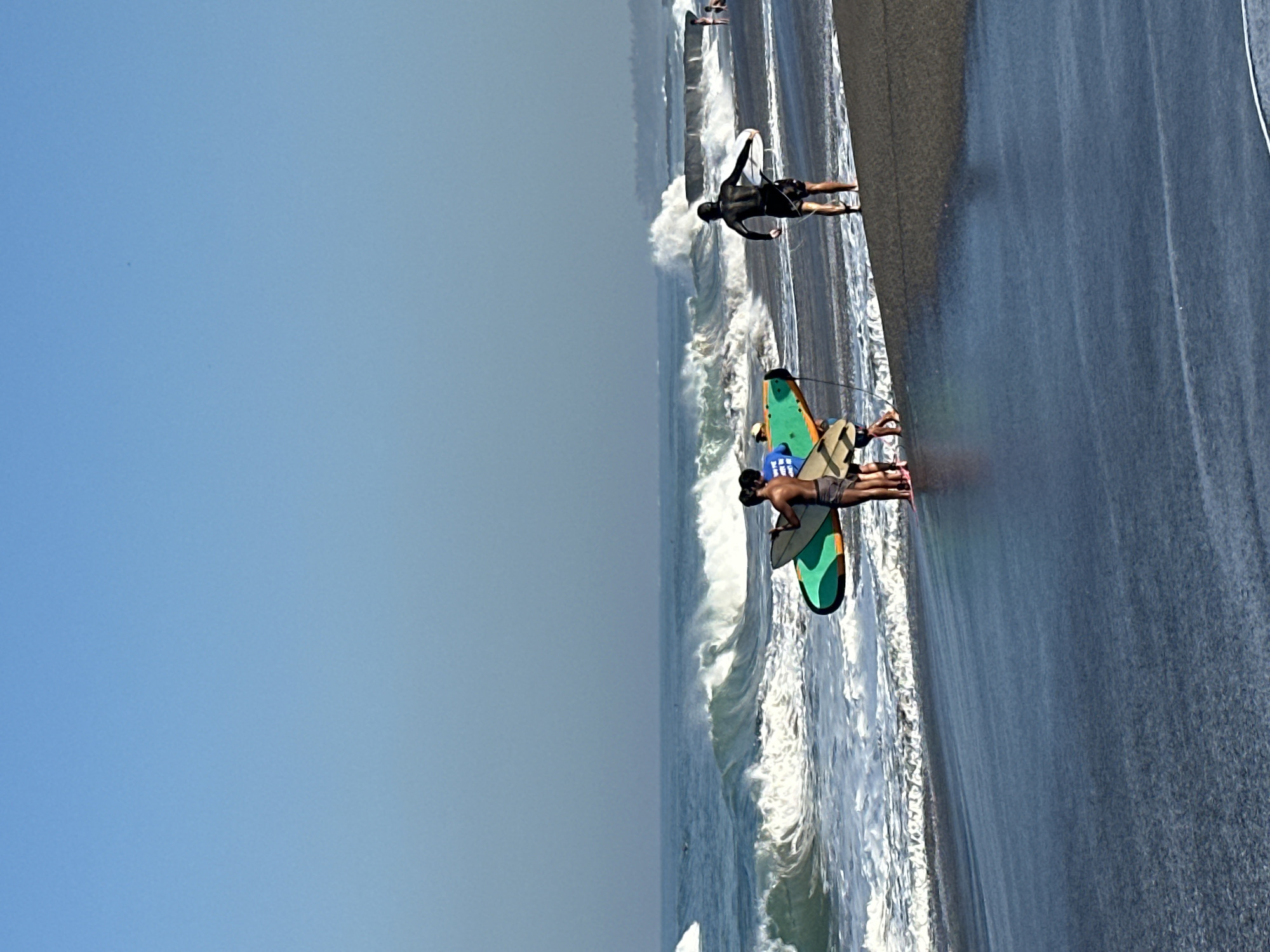 This is a photograph taken at Double Six Beach, North Kuta, Bali, Indonesia. The image depicts a beachfront scene with several individuals carrying surfboards. In the foreground, the dark sand beach is wet from the receding tide, with gentle waves lapping at the shore. A group of three people, two of whom are carrying a large green and orange surfboard, stand near the water's edge. To the right, a lone figure in a black wetsuit walks away from the camera, also carrying a surfboard. The ocean is a deep blue, with prominent white waves crashing and breaking further out. In the background, the horizon is hazy, with a faint outline of the coastline and what appears to be a breakwater or pier. The sky above is a clear, pale blue.