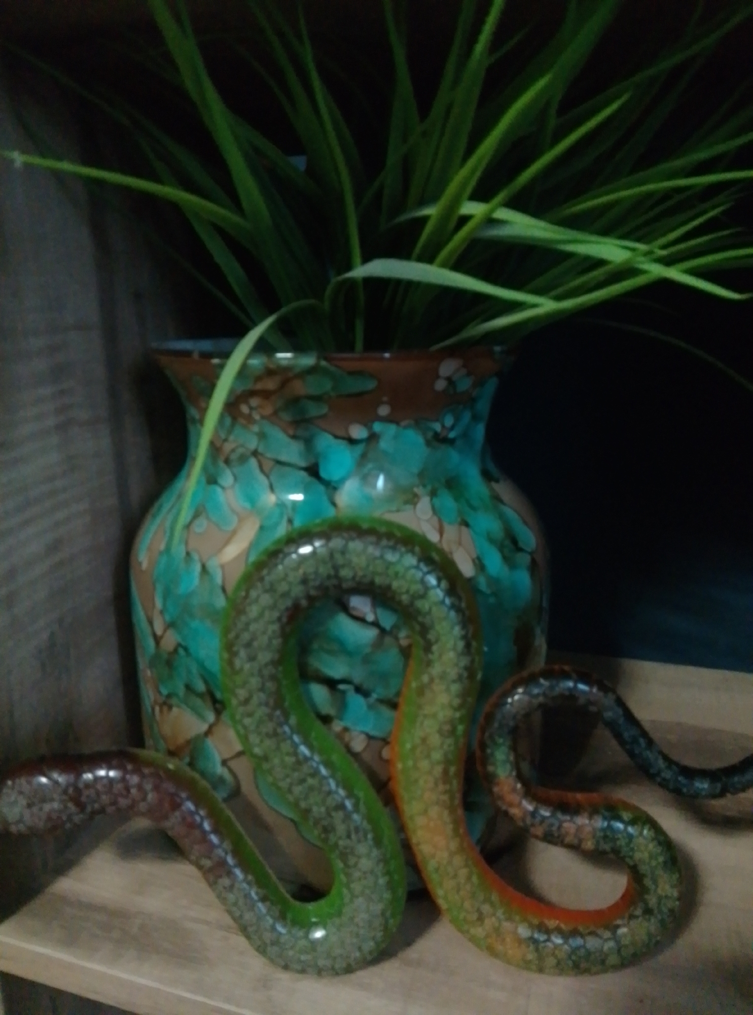 The image shows a decorative ceramic snake figurine positioned in front of a vase with a green and brown marbled pattern. The snake is coiled and its body is textured to resemble scales. The vase contains a lush green plant with long, thin leaves. The objects are placed on a wooden surface. The background is dark and out of focus. The image was taken inside a room, likely in Wedel, Germany.