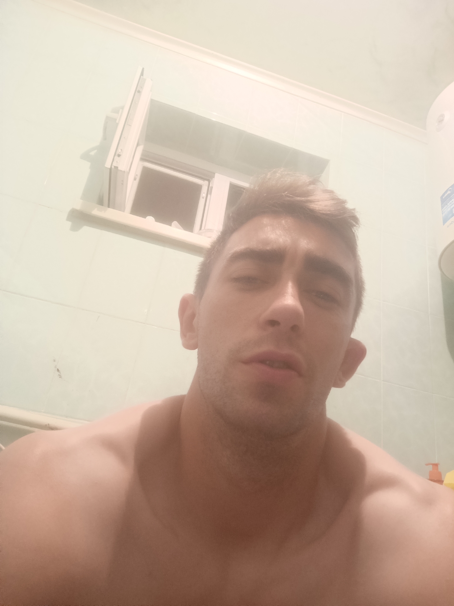 The image is a selfie of a young man, shirtless, in a bathroom. He has light brown hair and is looking directly at the camera with a neutral expression. The bathroom walls are tiled in a light greenish-blue color. A small, open window is visible in the background, with what appears to be a radiator below it. To the right of the man, a white cylindrical object, possibly a water heater, is mounted on the wall. The overall lighting is somewhat dim, suggesting it may be indoors or at dusk. There are no specific indicators in the image to suggest the location is in Fastiv, Ukraine.