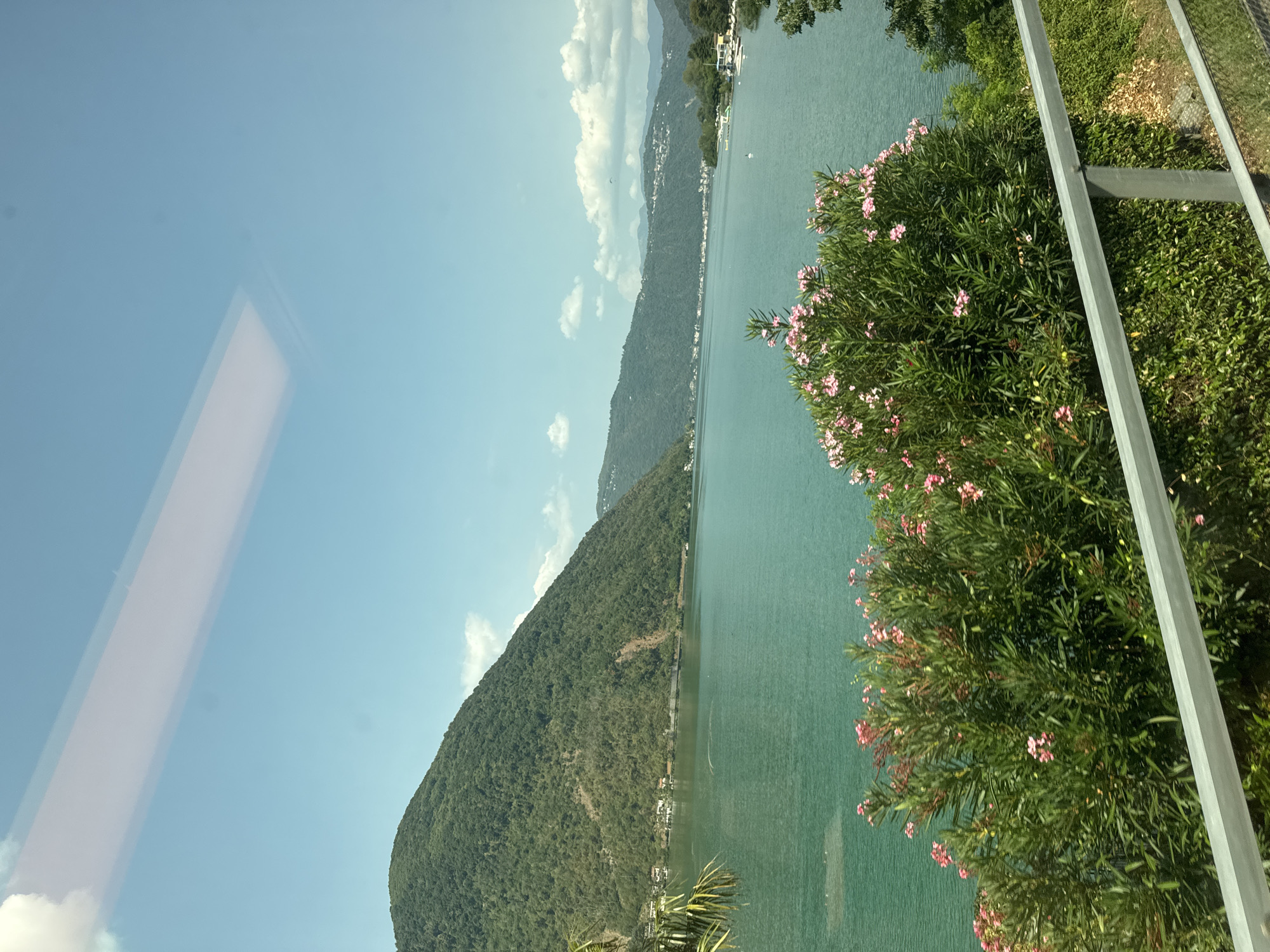 The image depicts a serene lakeside landscape under a clear blue sky with scattered clouds. In the foreground, a railing runs horizontally across the bottom, separating the viewer from lush green foliage and a flowering bush with pink blossoms. The middle ground is dominated by a vast expanse of tranquil turquoise water, likely a lake, reflecting the sky. Along the far shore of the lake, a verdant, tree-covered mountain rises steeply. Distant hills, also covered in vegetation and dotted with buildings, form a hazy backdrop. The overall impression is one of natural beauty and peacefulness. This scene is consistent with views from the area of Val Mara, Switzerland, which is situated on the shores of Lake Lugano and surrounded by mountains.