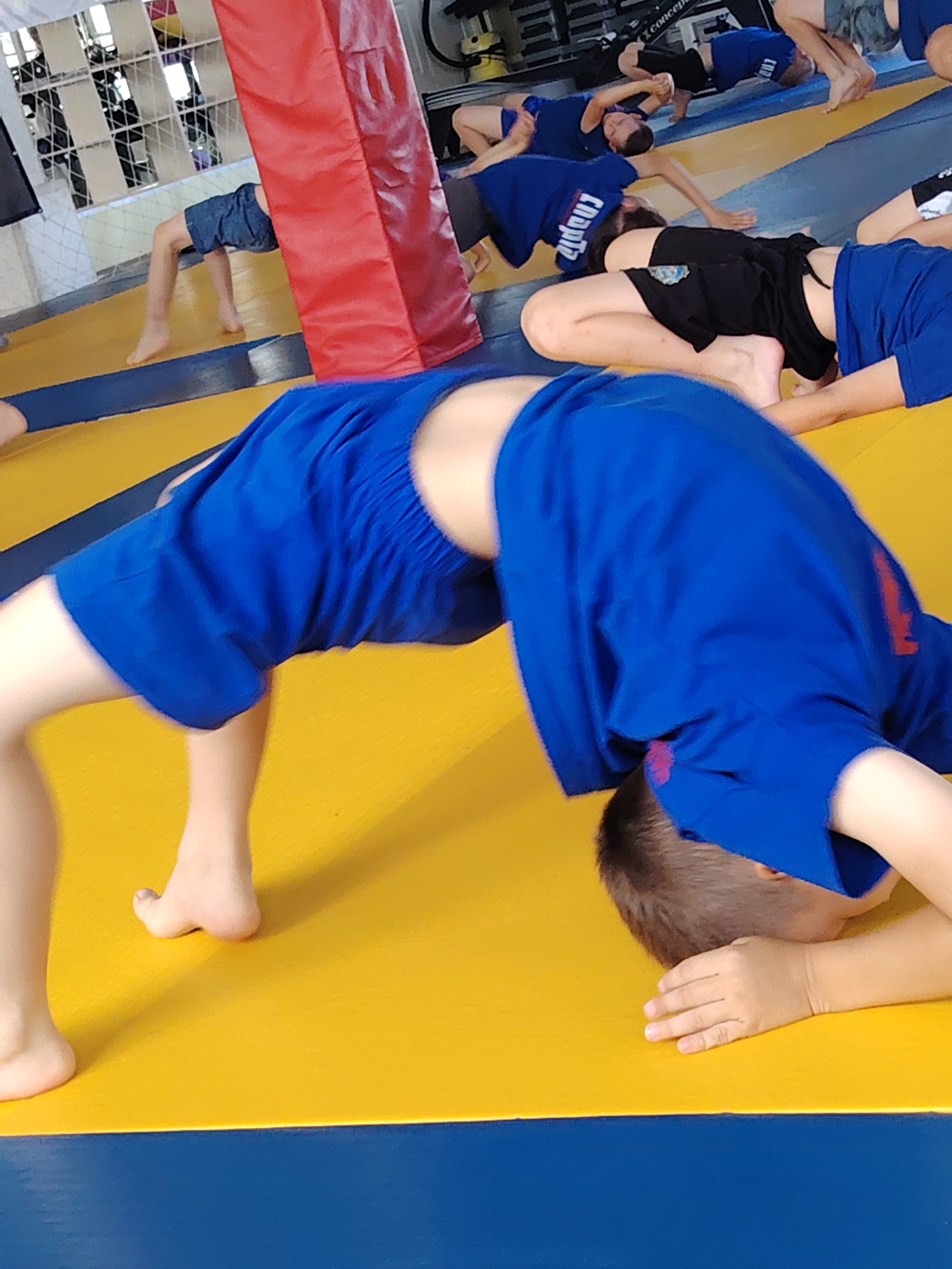 The image shows a group of children in a gymnasium, engaged in a physical training session. The gymnasium floor is a two-toned mat, primarily yellow with blue accents along the edge. In the foreground, a child is performing a backbend, with their body arched upwards, supported by their hands and feet. The child is wearing a blue uniform with red trim. Other children are visible in the background, some also in blue uniforms, lying on the mat or in similar stretching poses. A large, red, padded pillar is positioned to the left of the main subject, partially obscuring some of the background. The overall environment suggests a sports or martial arts training facility. Based on the provided information, this scene is located in a gymnasium in the city of Chaikovsky, Russia.