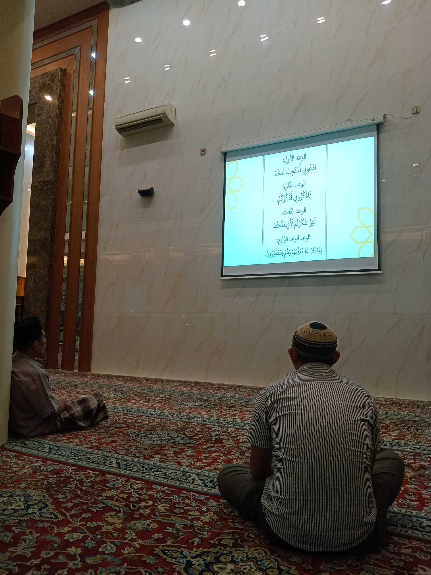 In a room with marble walls and a patterned carpet, two men sit facing a projector screen. The room appears to be a prayer room or mosque, with an air conditioning unit mounted on the wall. The projector screen displays Arabic text. The man on the left is wearing a brown shirt and a checkered sarong, while the man on the right is wearing a striped shirt and dark pants, with a patterned cap on his head. The man on the right is closer to the camera, with his back to the viewer, while the man on the left is further away and partially visible. The overall scene is dimly lit, with light emanating from the projector screen and overhead lights.