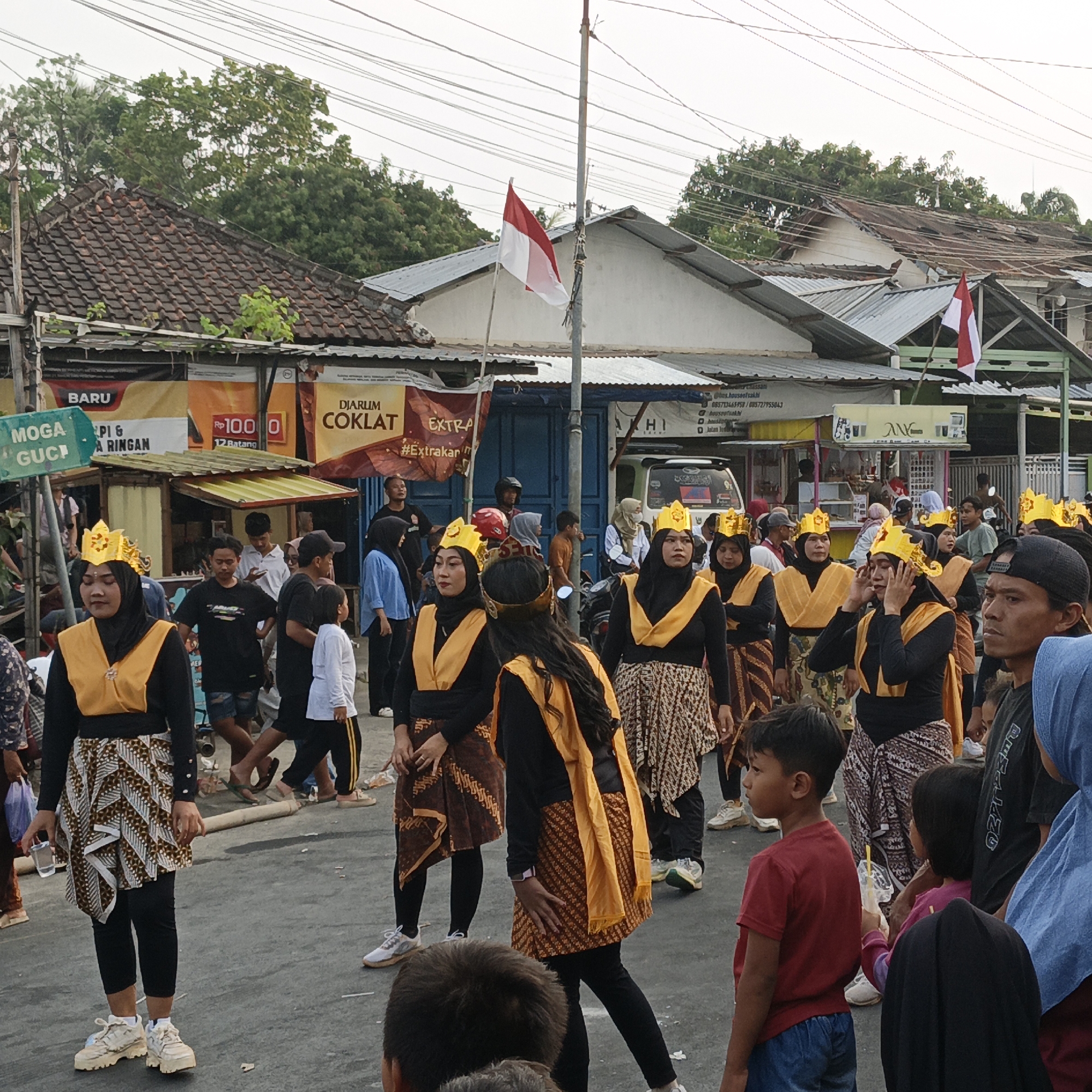 In Randudongkal, Indonesia, a group of individuals, predominantly women wearing traditional Indonesian attire including gold crowns and patterned skirts, are gathered on a paved street. Several of them are holding hands or are in close proximity, suggesting a coordinated activity or performance. Behind them, a row of small shops and market stalls are visible, with banners and signs indicating commercial activity. A flagpole with the Indonesian flag is also present. The sky is overcast, and the overall atmosphere appears to be that of a local festival or event.
