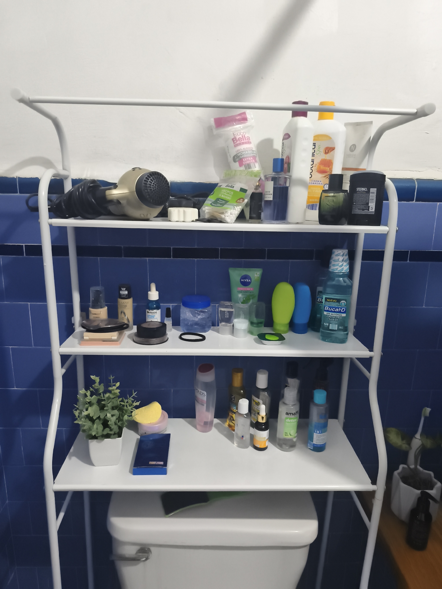 The image shows a white, four-shelf bathroom storage unit filled with various personal care items. The unit is positioned behind a white toilet in front of a wall partially tiled with blue square tiles.
On the top shelf, there's a gold-colored hairdryer, a box of cotton swabs, a bag of cotton pads, and several bottles and containers including lotion and fragrance.
The second shelf contains makeup items, face creams (Nivea), small tubes, bottles, and a bottle of Nuevo Bucal-D mouthwash.
The third shelf displays a small potted plant with a yellow sponge resting on it, several bottles and containers of skincare products (Pond's), including Amalfi detox and another bottle of 3 in 1.
The bottom shelf sits atop the toilet tank.
The setting appears to be a bathroom in Mexico City, Mexico. The location is inferred from some product labels.
