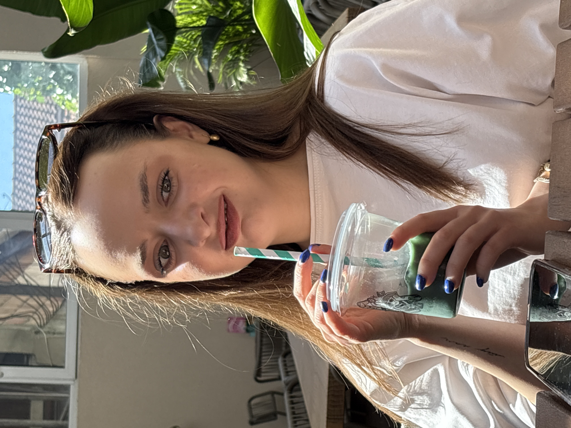 A young woman with long brown hair and blue nail polish is drinking a green beverage from a clear plastic cup with a green and white striped straw. She is wearing a white t-shirt and gold earrings. Sunglasses are perched on top of her head. The background suggests an outdoor cafe or restaurant setting with wooden tables and chairs, and lush green plants visible through a window. The lighting indicates it is daytime, possibly late morning or early afternoon. The scene appears to be in Kragujevac, Serbia, judging by the details provided, though specific landmarks are not identifiable.