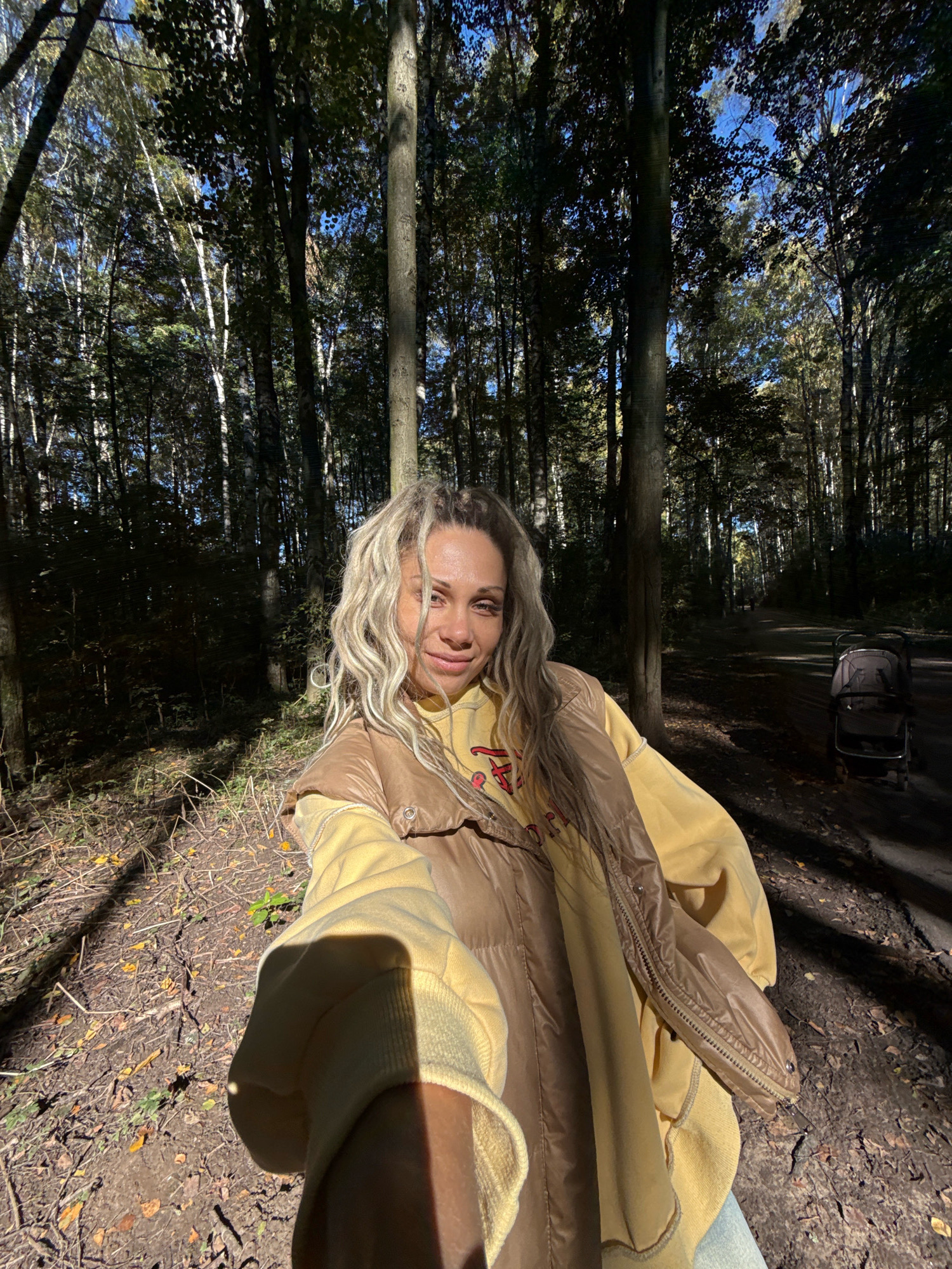 The image is a selfie taken in a wooded area, likely a park, given the presence of a path and a stroller in the background. A woman with blonde, wavy hair is in the foreground, smiling at the camera. She is wearing a yellow hoodie and a brown puffer vest. The ground is covered in dry leaves and twigs, suggesting an autumn setting. Sunlight filters through the trees, creating dappled shadows. The location is identified as Rusanovo, Russia, which is a city. A stroller is visible on the path to the right, and other people are faintly visible further down the path. The overall scene is outdoors, in a natural environment.