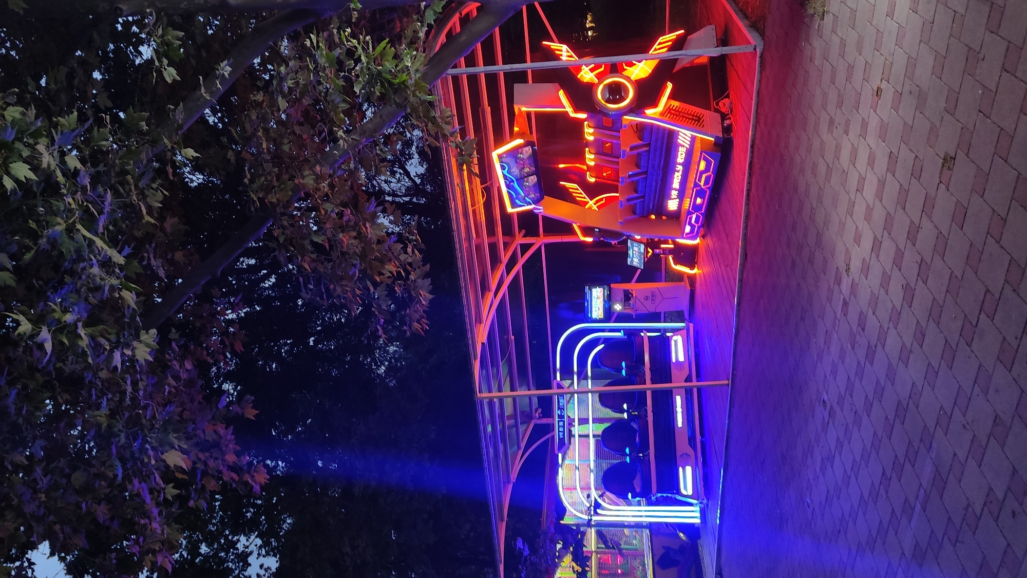 In the city of Krasnodar, Russia, at night, under the canopy of trees with leaves illuminated by neon lights, two virtual reality (VR) rides are set up outdoors. The ride on the left features a white structure with four seats, outlined in vibrant blue neon lights. Adjacent to it stands a control panel. To the right, a larger VR ride boasts a complex structure with multiple seats, predominantly illuminated by orange and red neon lights. This ride also has a screen displaying images of what appears to be virtual reality content. The ground is paved with interlocking bricks, reflecting the colorful lights of the rides. The overall atmosphere suggests an amusement park or entertainment venue at dusk or nighttime.