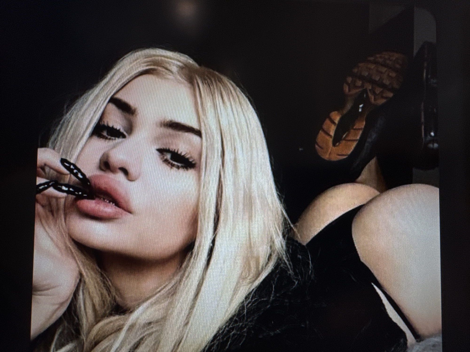 A close-up portrait shot features a person, likely female, with long, straight blonde hair framing her face. She has prominent dark eyebrows, heavily made-up eyes with defined winged eyeliner and long, dark eyelashes, and full, glossy lips. A distinctive black, pointed, claw-like object is held gently between her lips, adding a stylized or dramatic element to her pose. Her left hand is partially visible, supporting her chin.
The subject is dressed in dark, possibly black, attire that covers her upper body. In the upper right quadrant of the image, partially obscured, is a shoe or boot with a textured, light brown sole and a dark upper, appearing to be angled with the sole facing upwards. Below this, towards the right, a bare, light-skinned thigh or hip emerges from dark fabric.
The background is dark and indistinct, suggesting an indoor setting with low lighting or a dark backdrop. The lighting on the subject's face is soft, highlighting her features. The person maintains a direct and intense gaze towards the viewer, conveying a confident or alluring expression. The image itself appears to be a photograph of a digital screen, with visible scan lines or pixelation present across the composition. No text is visible in the image, and there are no specific visual cues to indicate the stated location of Varna, Bulgaria.