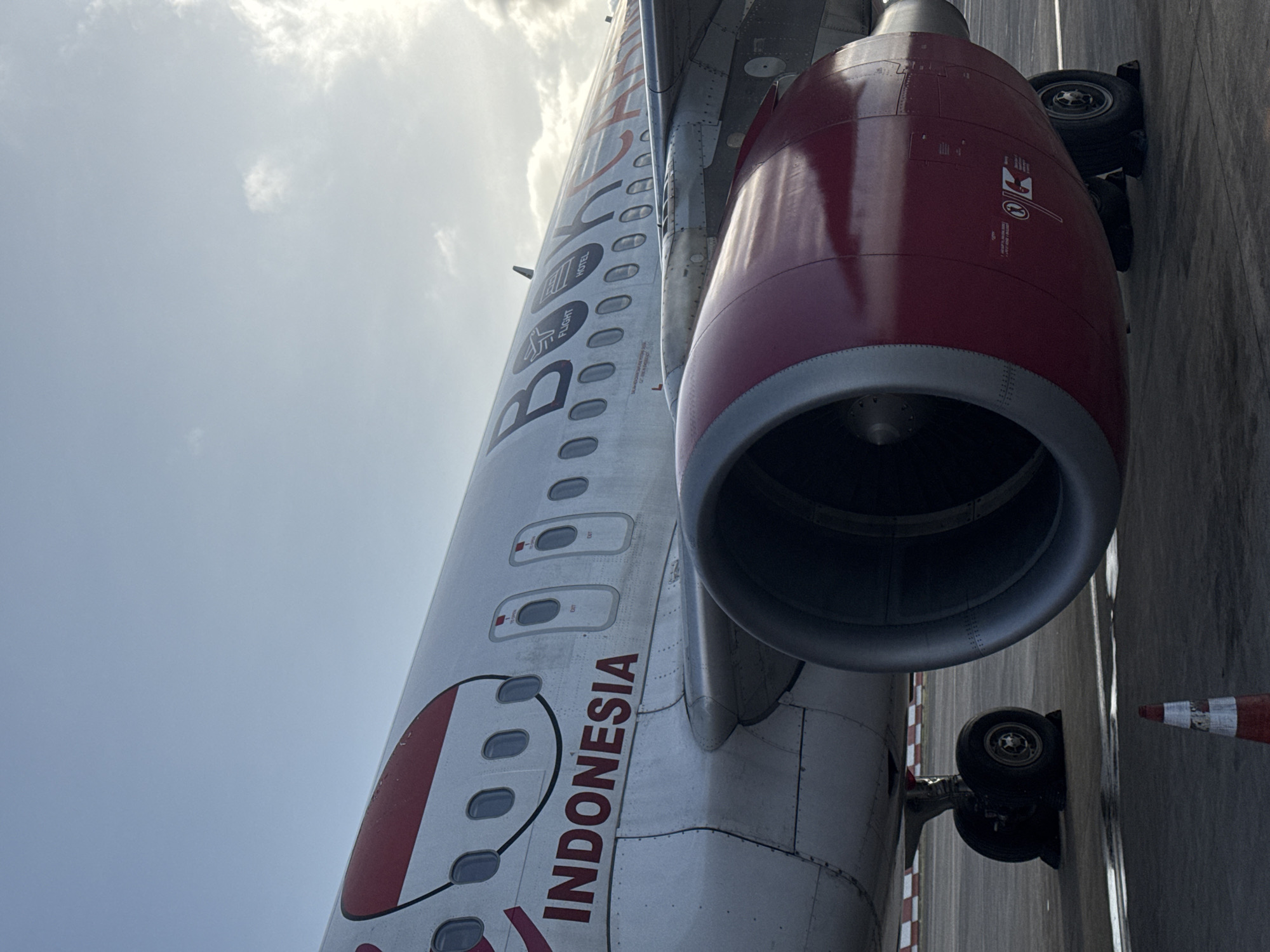 A close-up, angled shot of an airplane fuselage on a tarmac, with a prominent red engine casing dominating the right side of the frame. The fuselage is painted white with red accents, and "INDONESIA" is clearly visible in red lettering. The word "BATIK" is also distinguishable above "INDONESIA". A row of oval windows is present along the fuselage. The plane's landing gear is visible beneath the engine. A partially visible orange and white cone is positioned on the ground to the right. The background features a bright, cloudy sky. The location is the city of Unknown, Unknown.