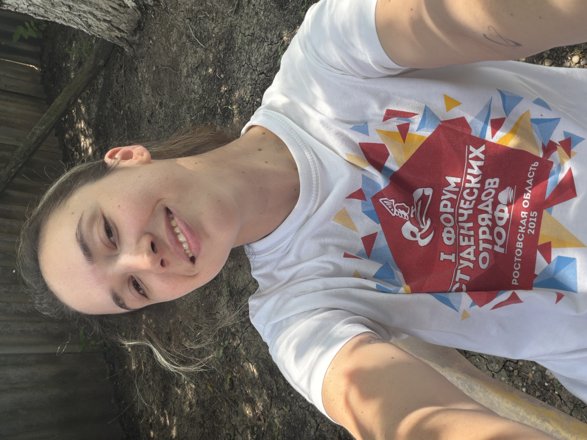 A young woman with light brown hair pulled back smiles for a selfie. She is wearing a white t-shirt with a red, yellow, and blue graphic design and text. The text on the shirt reads "I ФОРУМ СТУДЕНЧЕСКИХ ОТРЯДОВ ЮФО РОСТОВСКАЯ ОБЛАСТЬ 2015". She is seated outdoors in what appears to be a yard, with dry, cracked ground and a wooden fence visible in the background. A tree trunk is also present to the right. The lighting suggests it is daytime.