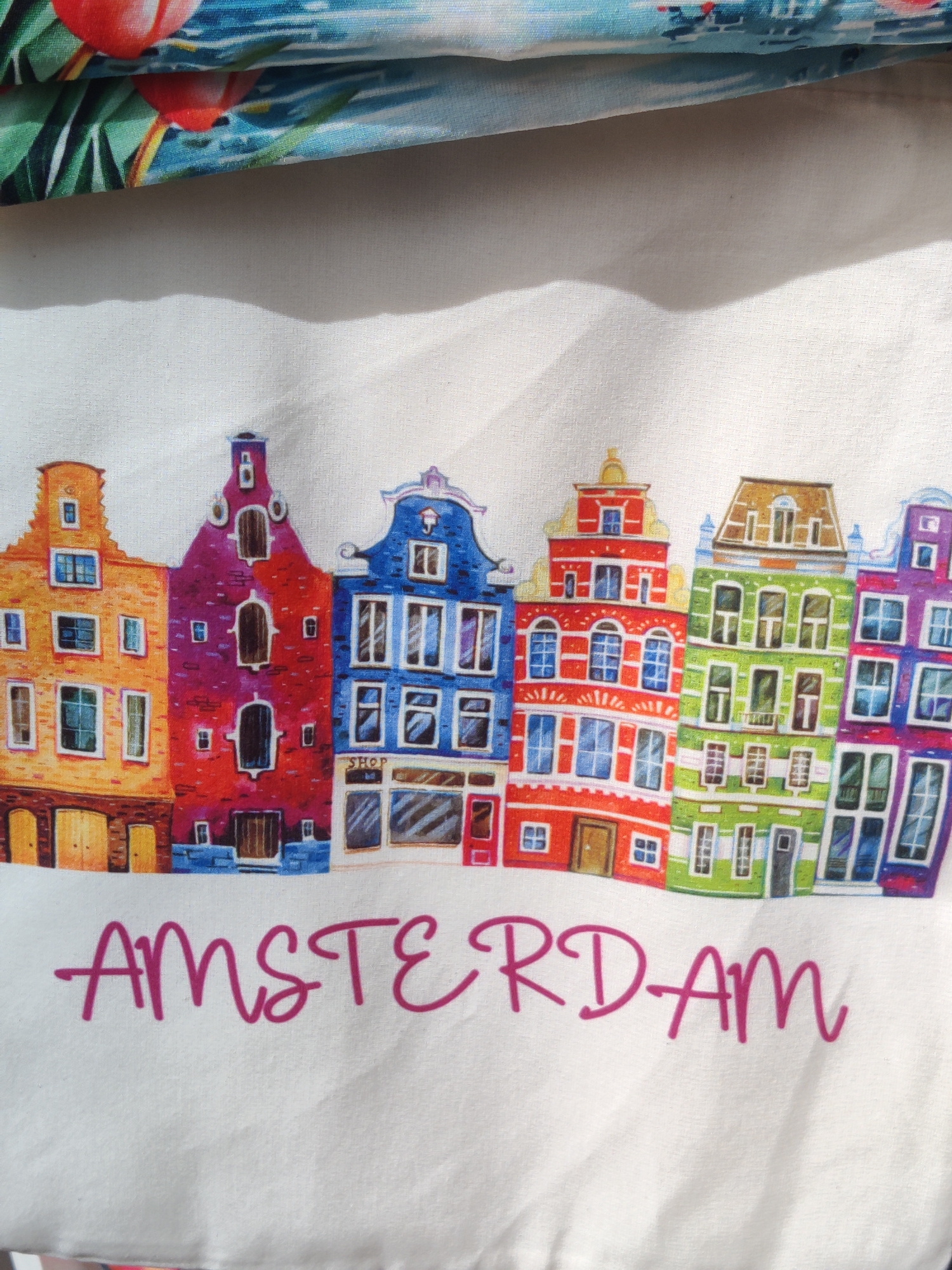 The image shows a close-up of a white fabric, possibly a tote bag or a tea towel, featuring a stylized depiction of Amsterdam canal houses in various vibrant colors. The houses are arranged in a row, each with distinct architectural features like gabled roofs and arched windows. The word "AMSTERDAM" is printed in large, magenta script across the bottom of the fabric. Above the houses, a blurred image of tropical leaves and red flowers can be seen, suggesting a decorative border or another part of the fabric design. The overall impression is a charming and colorful souvenir item related to Amsterdam.