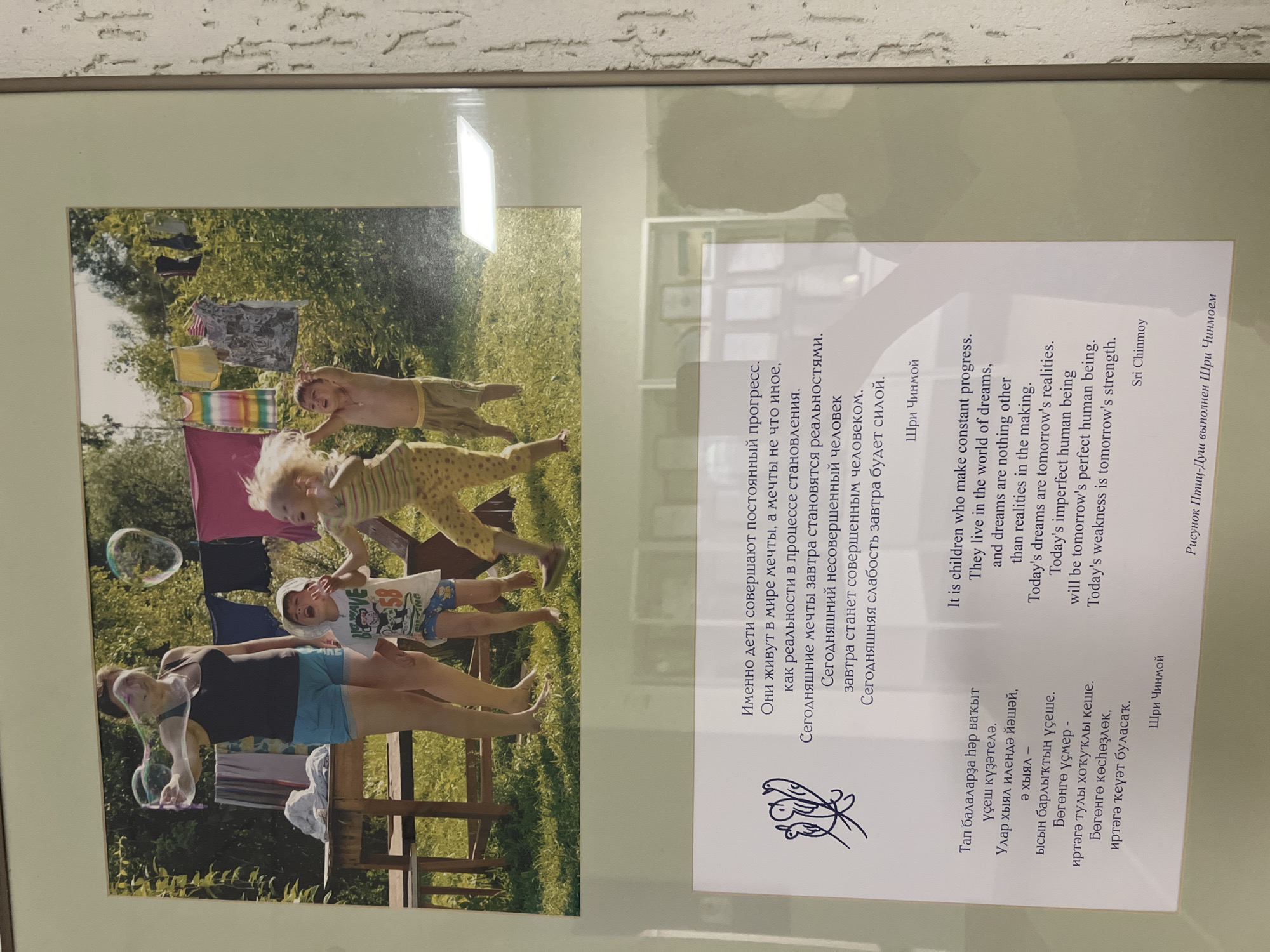 The image shows a framed print. The print contains a photograph of a woman and three children outside. The woman is blowing bubbles while lying on a wooden platform or bench. The children are interacting with the bubbles. Laundry is hanging on a line in the background, with trees visible beyond the laundry.
The right side of the print includes text in three languages (likely Russian, English, and another). The text is attributed to Sri Chinmoy. There is also a small butterfly-like symbol below the Russian text. The text is about children making constant progress and how dreams become realities. At the very bottom right corner, it mentions "Рисунок Птиц-Душ выполнен Шри Чинмоем".
The location of Sanatoriya Yumatovo Imeni 15-Letiya Bassr, Russia is not discernable from the image alone.