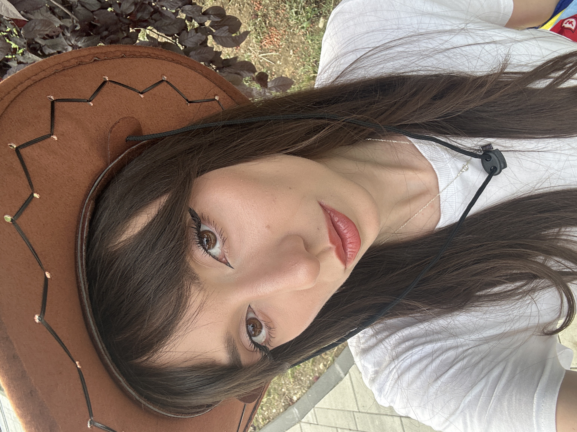 In an outdoor, close-up portrait, a young woman with long, dark brown hair and bangs wears a brown cowboy hat and a white t-shirt. Her makeup includes eyeliner and lipstick. A silver necklace is visible. The background shows dark foliage and a paved walkway. The image appears to have been taken in the city of Grocka Urban Municipality, Serbia.