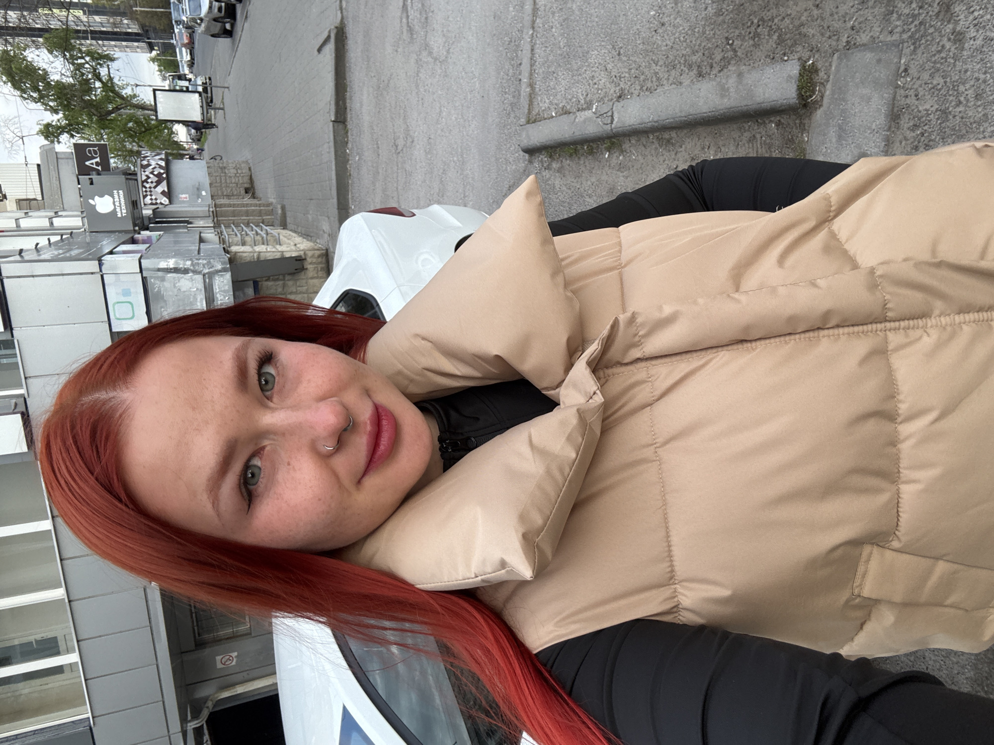 A medium close-up outdoor portrait features a young woman with long, reddish-orange hair and light green eyes, looking directly at the camera with a gentle smile. She has freckles across her nose and cheeks and wears a silver nose ring. Her head is tilted slightly to her left shoulder. She is dressed in a light beige or tan puffer vest over a black long-sleeved top.
The background shows an urban street scene in Rostov-on-Don, Russia. To the woman's left, partially visible, is a white car. Beyond the car and the woman, a grey building with multiple windows and balconies can be seen. Further back, along what appears to be a sidewalk or pedestrian area, there are various signs and structures. One prominent sign features an Apple logo and the Russian text "МАГАЗИН ТЕХНИКИ," which translates to "ELECTRONICS STORE." Another sign above it displays "Aa." In the distance, other parked cars line the street, and a bare-branched tree suggests cooler weather or an early spring day. The sky appears overcast, indicating daytime with muted lighting. The overall impression is a casual outdoor snapshot taken on a city street.