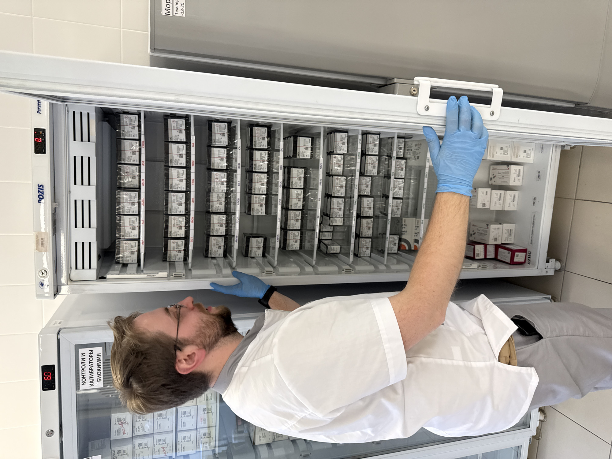 A man with a beard and glasses, wearing a white lab coat over a grey shirt and blue protective gloves, is reaching into a large, white laboratory refrigerator. He is holding the top handle with his right hand, while his left hand is inside, appearing to organize or retrieve items.
The refrigerator is packed with numerous small, rectangular boxes, likely reagent kits or laboratory supplies, neatly arranged on multiple shelves. Labels on the boxes indicate various biochemical assays and analytes, such as CHOL, AST, ASO, GLUC, LDL, HDL, FERR, NACL, CYSC, МЕДЬ (Copper), цинк (Zinc), GLDH, TRG, LDH, IRON, UA, TRSF, GGT, ALP, AMYL, CRPHS, A2MG, ALB, BILT, APOB, CREA, UREA, CHE, PHOS, CA, MG, GLU.
On the left side of the refrigerator, a digital display shows "6.0". Below it, a label reads "КОНТРОЛИ И КАЛИБРАТОРЫ БИОХИМИЯ" (CONTROLS AND CALIBRATORS BIOCHEMISTRY). Another digital display higher up shows "8.8.", and the brand names "POZIS" and "Paracel" are visible. Above this unit, a second refrigerator or freezer is partially visible, with text "Морг" (Morgue – likely a brand or model name), "Темпер" (Temperature), and "-18-20", indicating a freezer. The setting is a laboratory or medical facility with white tiled walls, and the man appears focused on his task.