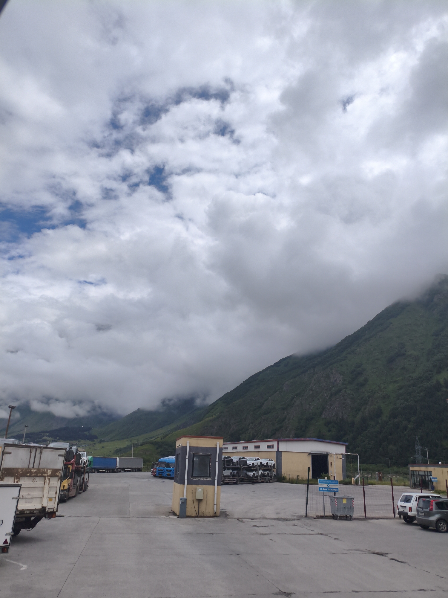 The image depicts an outdoor scene at what appears to be a truck depot or customs checkpoint, likely located in Achkhoti, Georgia, given the mountainous terrain. The sky is heavily laden with large, dramatic clouds, with patches of blue sky visible. In the foreground, several large trucks are parked on a paved surface. Some of these trucks are trailers, with one particularly prominent in the lower left, showing a weathered white and beige cargo container.
Behind the parked trucks, there's a low-rise building with a yellow facade and a dark open doorway. A truck with a trailer carrying multiple white cars is parked in front of this building. Further back, a bright blue truck is visible, along with other long-haul trucks. A small, enclosed booth with windows and a grey panel is positioned near the center of the image, suggesting a checkpoint or security station.
The background is dominated by a steep, green mountainside, with parts of the upper slopes shrouded in mist and clouds, indicating a high-altitude or rainy environment. The overall impression is one of a functional, industrial area set against a dramatic natural backdrop. There are no people visibly present in the scene.