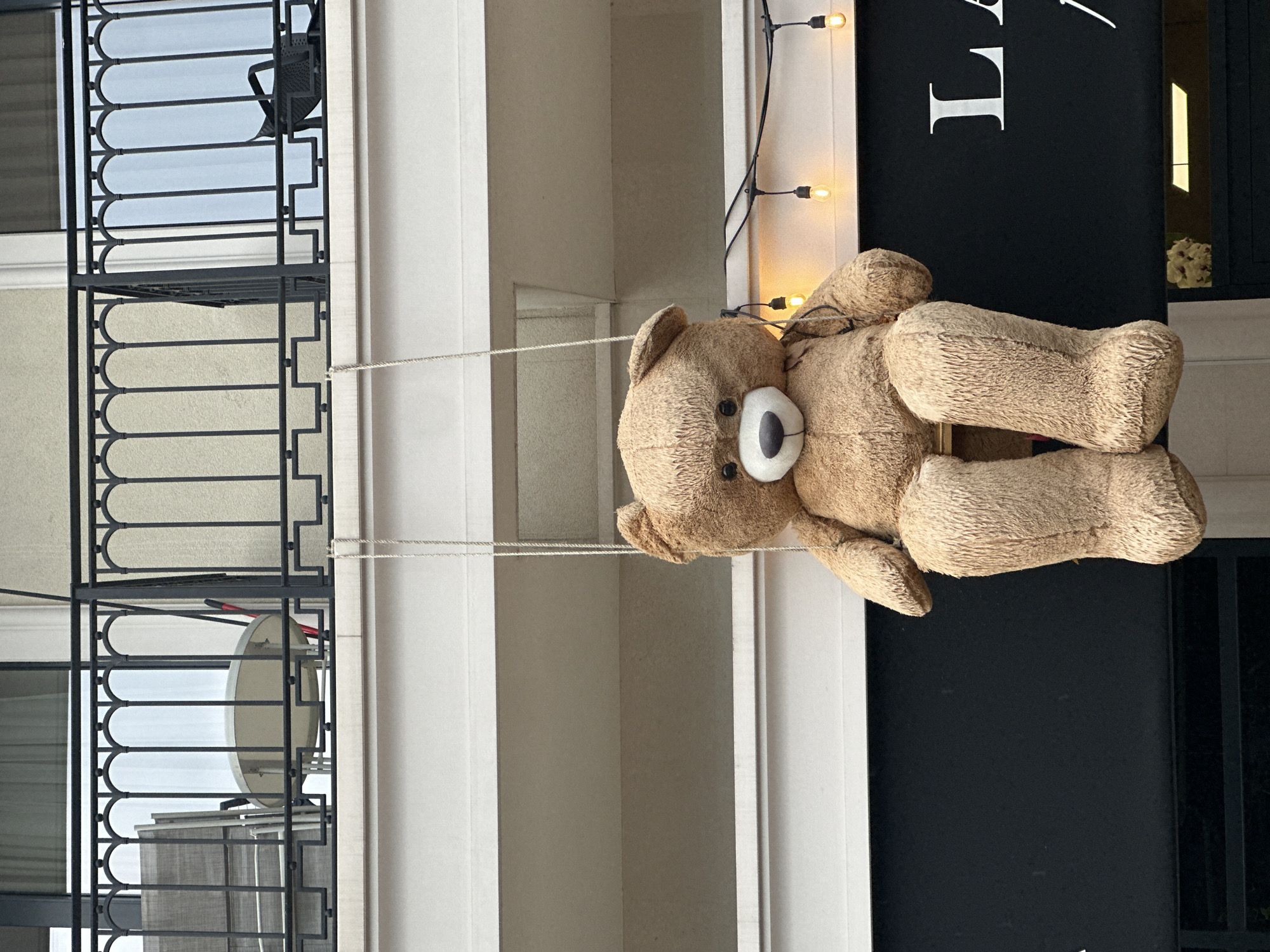 A large, light brown teddy bear hangs vertically as a decoration against a building facade. The bear, made of a shaggy, textured material, has a distinctive white snout and a black nose. It is suspended by two thin, light-colored wires or ropes, with its arms stretched upwards by the suspension.
To the bear's right, a prominent black sign features large white letters. The letter "L" is clearly visible, followed by what appears to be part of another letter, possibly an "A" or a similar character, and then a decorative swirl. Below this visible text, a string of warm, glowing lights is draped vertically, adding to the decorative ambiance.
To the bear's left, the building features several balconies with elaborate black wrought-iron railings. These railings display intricate patterns with arched top sections. Behind the railings, parts of windows and the light-colored building facade are visible. On one of the balconies, a large, round, light-colored object, possibly a satellite dish or a fan, is discernible. Below it, a dark mesh screen or fabric is also present. A glimpse of a red handle from an object can be seen on another balcony.
The scene suggests an outdoor decorative display, possibly for a shop or establishment, creating a welcoming and whimsical atmosphere. The presence of both daylight and glowing string lights implies either a daytime setting with decorative lights or a transitional time of day. While the exact location is not discernible from the image, it is situated in Budva, Montenegro, as per the provided context.