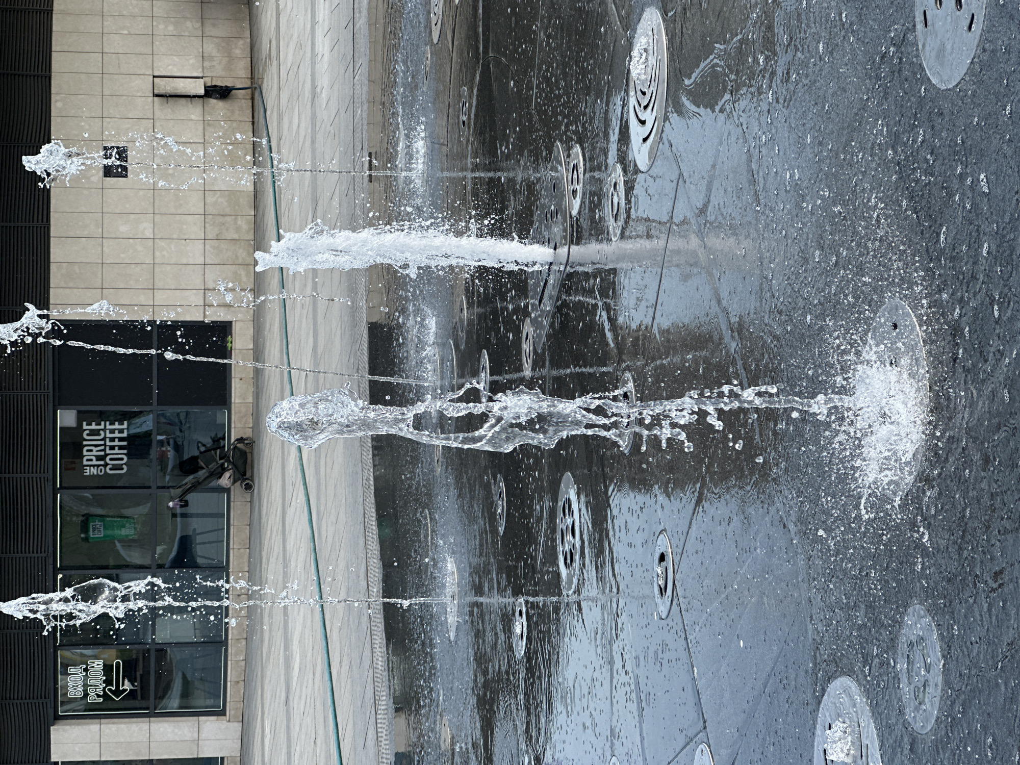 An outdoor fountain with multiple jets of water spraying upwards is centered in the image. The ground is a dark, possibly tiled, surface with metal grates or drain covers embedded within it. The reflection of the sky and surrounding buildings is visible on the wet surface.
To the left of the fountain, a building is partially visible. The facade appears to be made of light-colored stone or concrete. A storefront with dark-framed windows and the words "PRICE COFFEE" in English and "ВХОД РЯДОМ" (Entrance Next) in Russian are seen on one of the windows.
The photo was taken in Moscow, Russia.