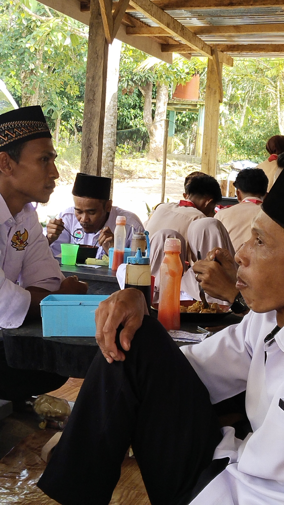 In Margatunggal, Indonesia, several men wearing black peci hats are seated at a table outdoors. The scene appears to be under a covered outdoor dining area with a corrugated metal roof supported by wooden beams. The men are dressed in white shirts, some with national emblems. They are engaged in eating and socializing. On the table, there are plates of food, bowls, plastic cups, and various sauce bottles, including red and brown condiments. The environment surrounding the dining area is lush with green trees and vegetation, indicating a rural or semi-rural setting. The lighting suggests it is daytime, with sunlight filtering through the trees. One man in the foreground is looking to the right, while others are engaged with their food or conversing. The overall atmosphere suggests a casual meal shared amongst a group.