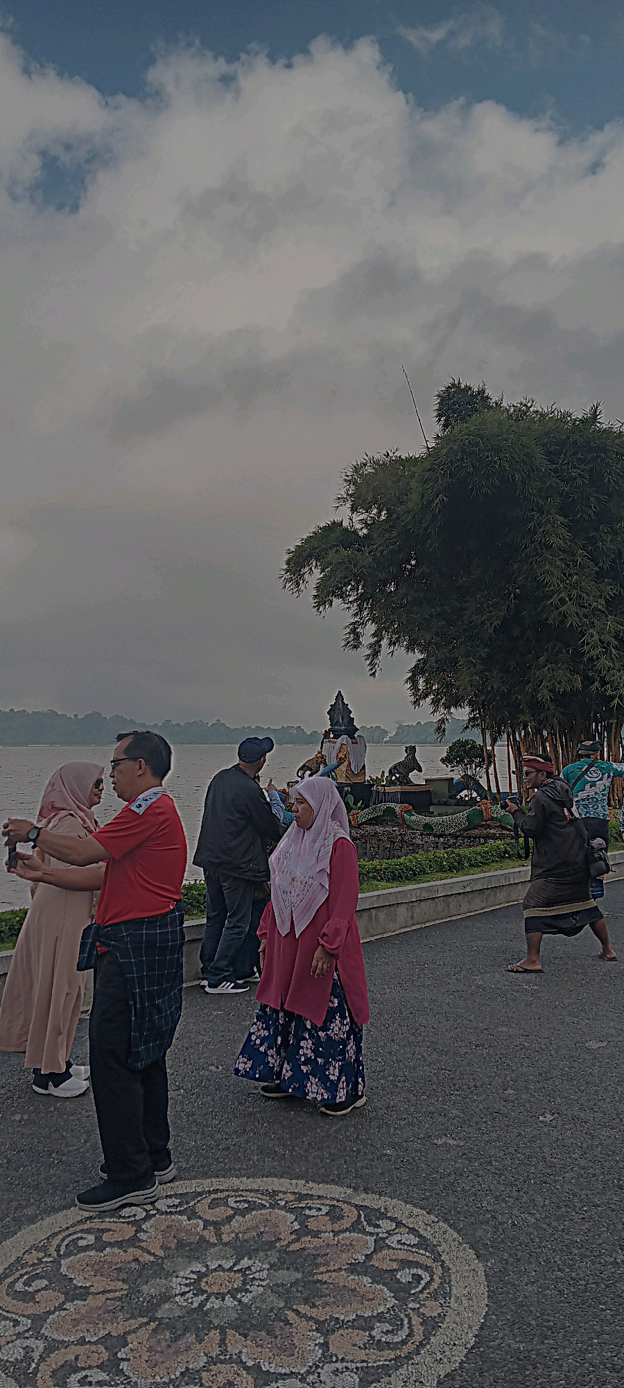 A group of people are standing by a body of water, possibly a lake or reservoir, near a decorative structure. The sky is overcast with clouds. In the foreground, a circular mosaic pattern is visible on the paved ground. On the left, a couple is taking a selfie, with the woman wearing a light-colored hijab and dress, and the man wearing a red shirt and plaid pants. To their right, another couple is visible, with the man wearing a dark jacket and baseball cap, and the woman wearing a pink hijab and a patterned skirt. Further to the right, a man in traditional clothing is walking with a camera around his neck. Behind the people, a bamboo tree provides shade. The decorative structure appears to be a shrine or monument, featuring sculptures of animals, possibly tigers and a dragon-like creature, with a central religious figure. The environment suggests a tourist area or scenic spot. The overall scene is set in the city of Baturiti, Indonesia.