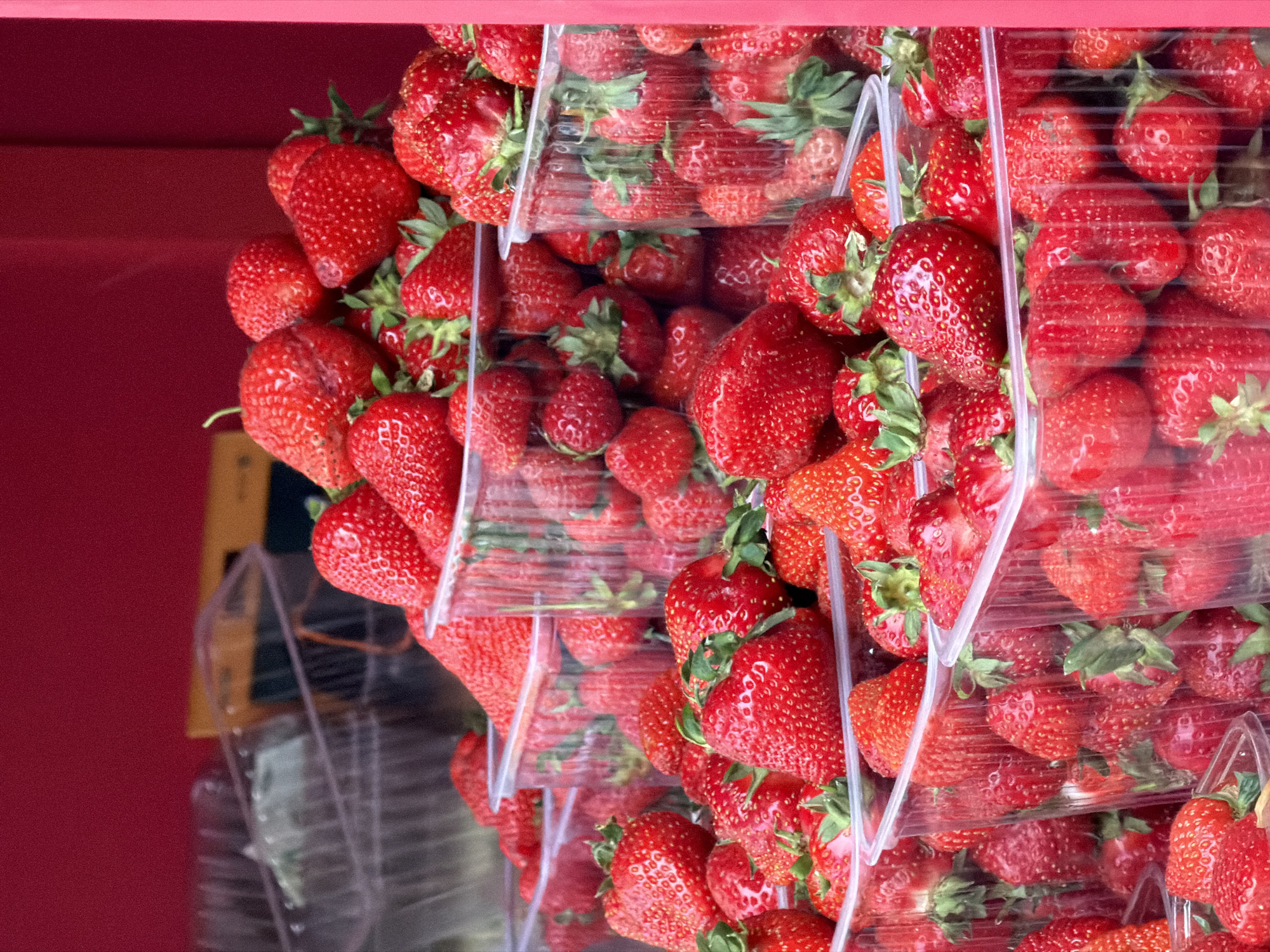 The image displays multiple containers of fresh strawberries stacked for sale, possibly at a market. The strawberries are a vibrant red color and appear to be plump and ripe. The containers are made of clear plastic, allowing the strawberries to be easily seen. The background is a solid red color, which might be the wall of a market stall or the side of a vehicle. There is no clear indication of the exact location in Moscow, Russia, or any information regarding people, interactions or specific actions within the image.