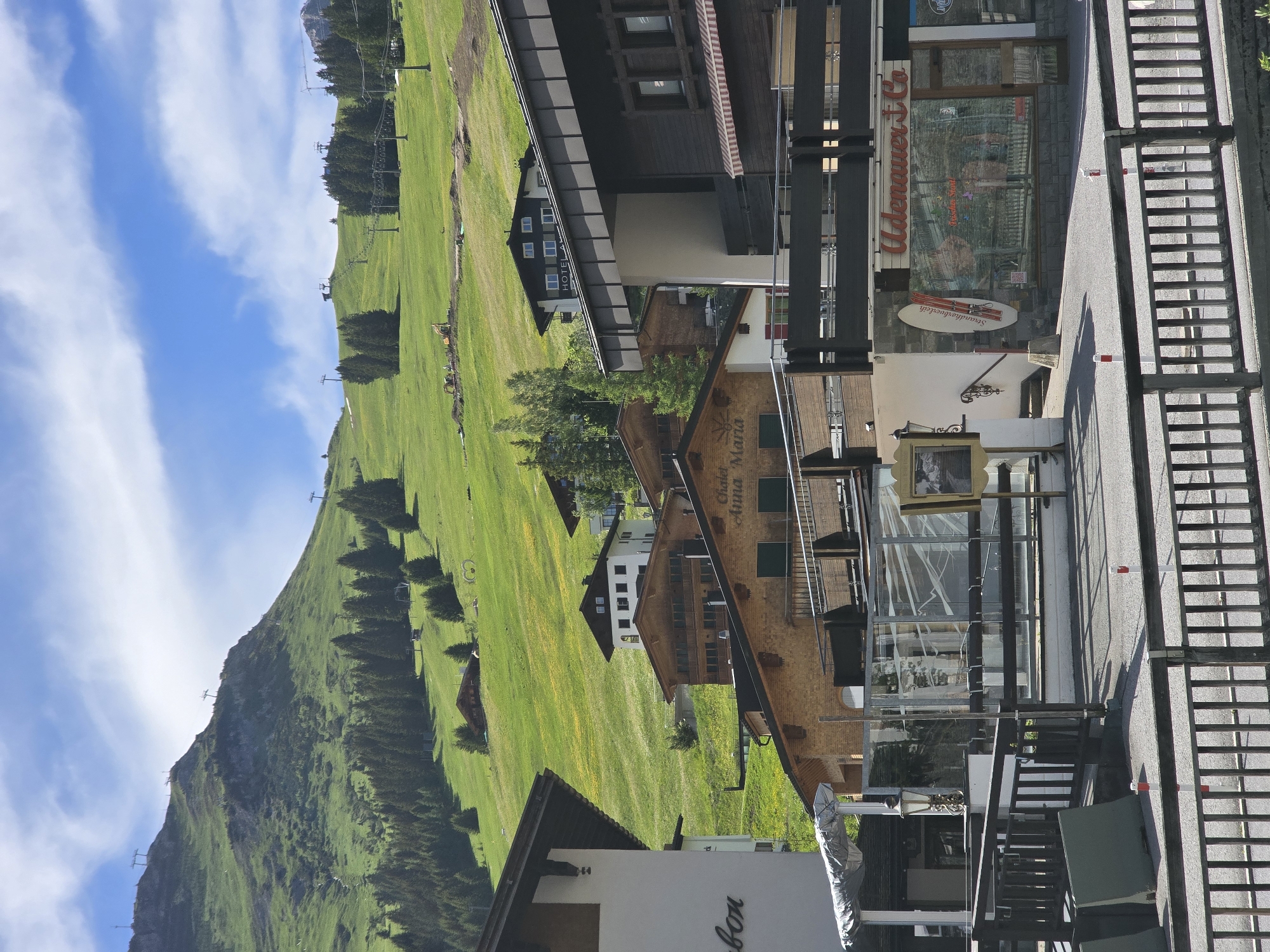 The image captures a view of a mountainside village, likely in Lech, Austria, on a sunny day with a partly cloudy sky. The foreground shows a street with a wooden fence and bollards. Buildings with traditional alpine architecture, featuring wood and stone facades, are visible. Some buildings have balconies and awnings. Signs on the buildings indicate businesses like "Chalet Anna Maria" and "Udenauer & Co," suggesting a commercial area.
In the background, a lush green mountainside slopes upwards, dotted with coniferous trees and ski lifts, indicating a ski resort. Some construction or excavation work is noticeable on the lower part of the mountain. The overall scene depicts a picturesque mountain village setting, likely during the summer season given the green landscape. There are no people visible in the image.