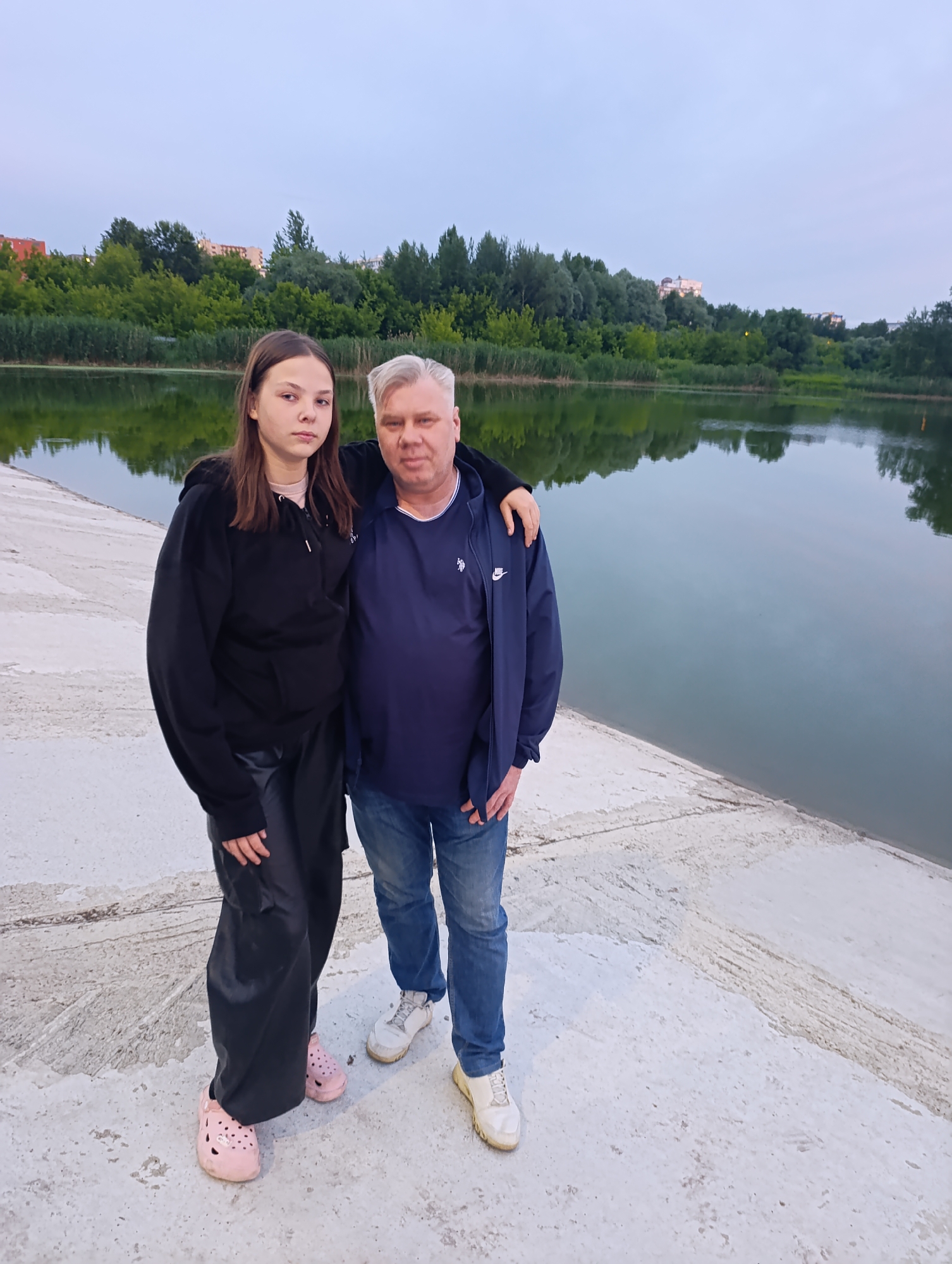 A full-shot outdoor portrait captures a middle-aged man and a young woman standing on a concrete embankment next to a large body of water. The man, positioned on the right, has fair skin, graying hair, and is wearing a dark blue Nike jacket over a blue t-shirt and blue jeans. He has his arm around the young woman. The young woman, on the left, has fair skin and is wearing a black hoodie, black pants, and pink Crocs. Her left arm rests on the man's arm. The water reflects the overcast sky and a line of trees and buildings across the bank. The concrete embankment shows signs of wear and tear, with visible lines and some discoloration. The overall atmosphere is subdued, likely due to the twilight or overcast weather. The location is likely a park or recreational area in Penza, Russia, given the context provided.