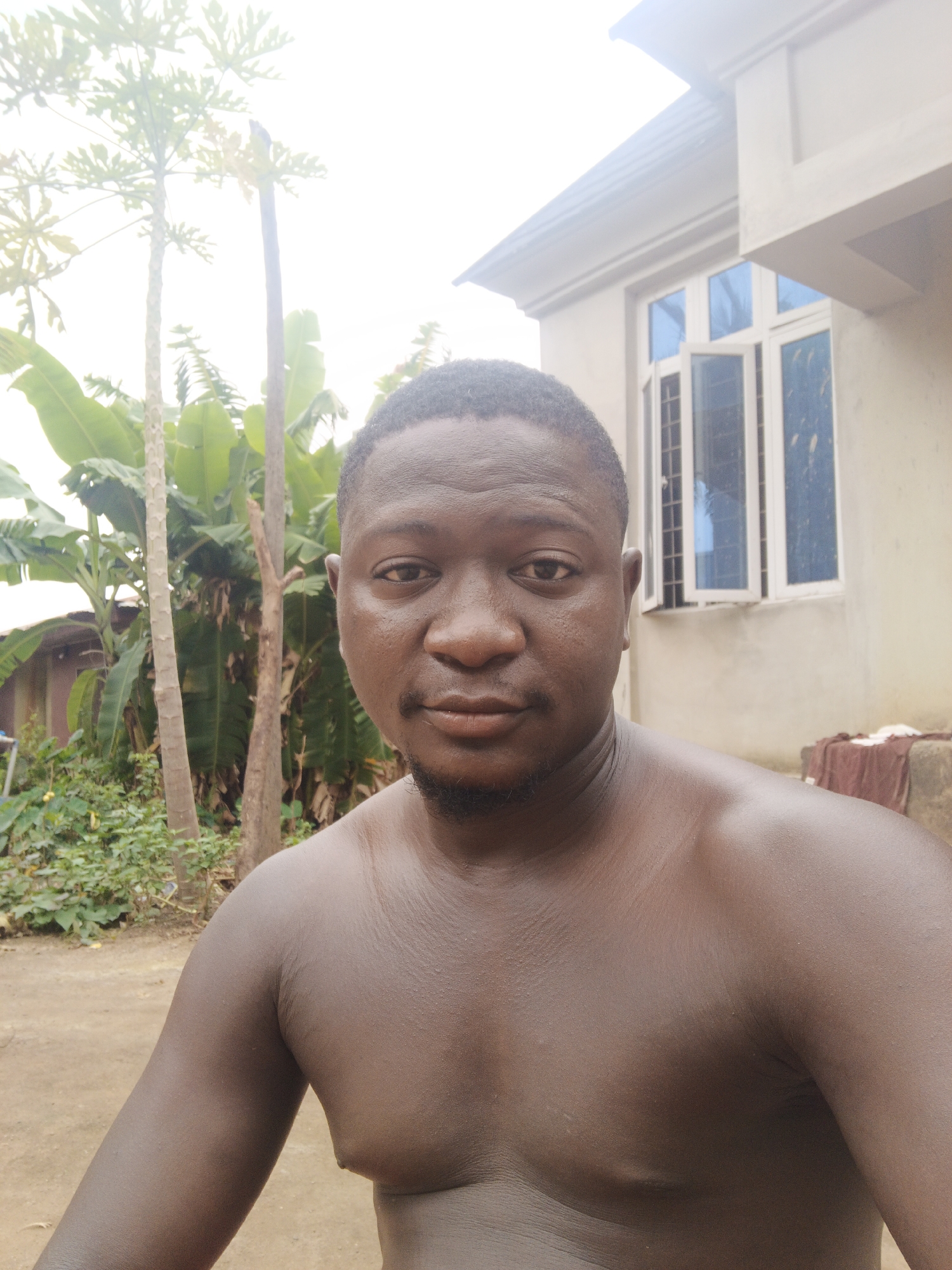 The image depicts a shirtless man with dark skin, facing the camera. He has short, dark hair and a small beard. The background features vegetation, including trees with broad leaves, and a building with white-framed windows. The location is likely Akwanga, Nigeria, given the prompt's context.