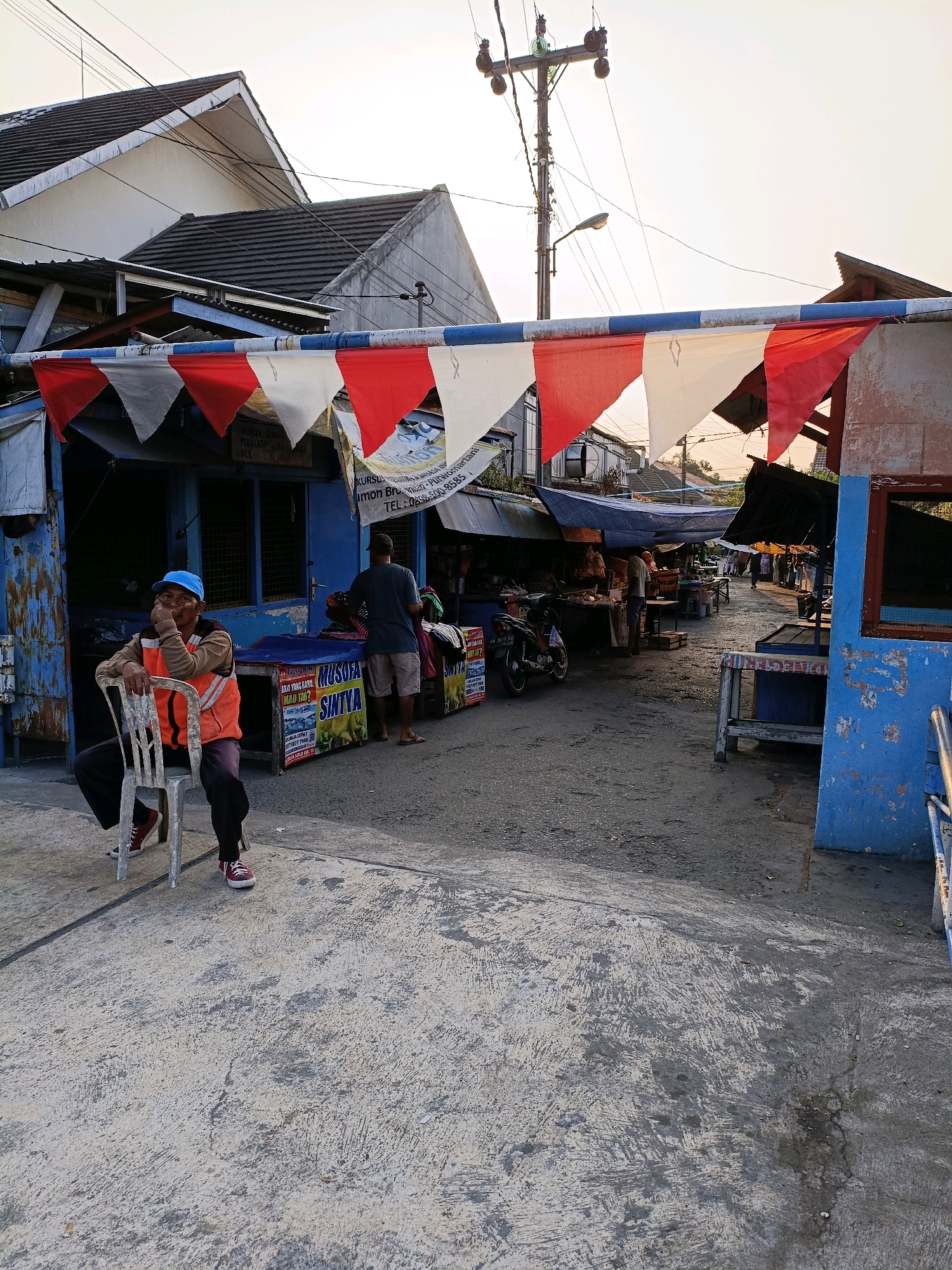 In Purwomartani, Indonesia, a man sits on a metal chair outside a market stall. He wears a blue cap, an orange safety vest over a brown long-sleeved shirt, and dark pants with red sneakers. The man is observed with his hand near his face, appearing to be contemplating or resting. Behind him, a blue market stall displays signage, including "MUSOFA SINTVA". The street is paved with asphalt and concrete. Other market stalls and awnings are visible, with several people browsing or working. A motorcycle is parked to the left of the scene. Red, white, and blue pennants are strung across an overhead wire, likely for decoration. The sky is a pale yellow, suggesting either early morning or late afternoon light. Electrical wires and a utility pole are prominent in the background, typical of urban infrastructure. The overall atmosphere is that of a bustling local market.