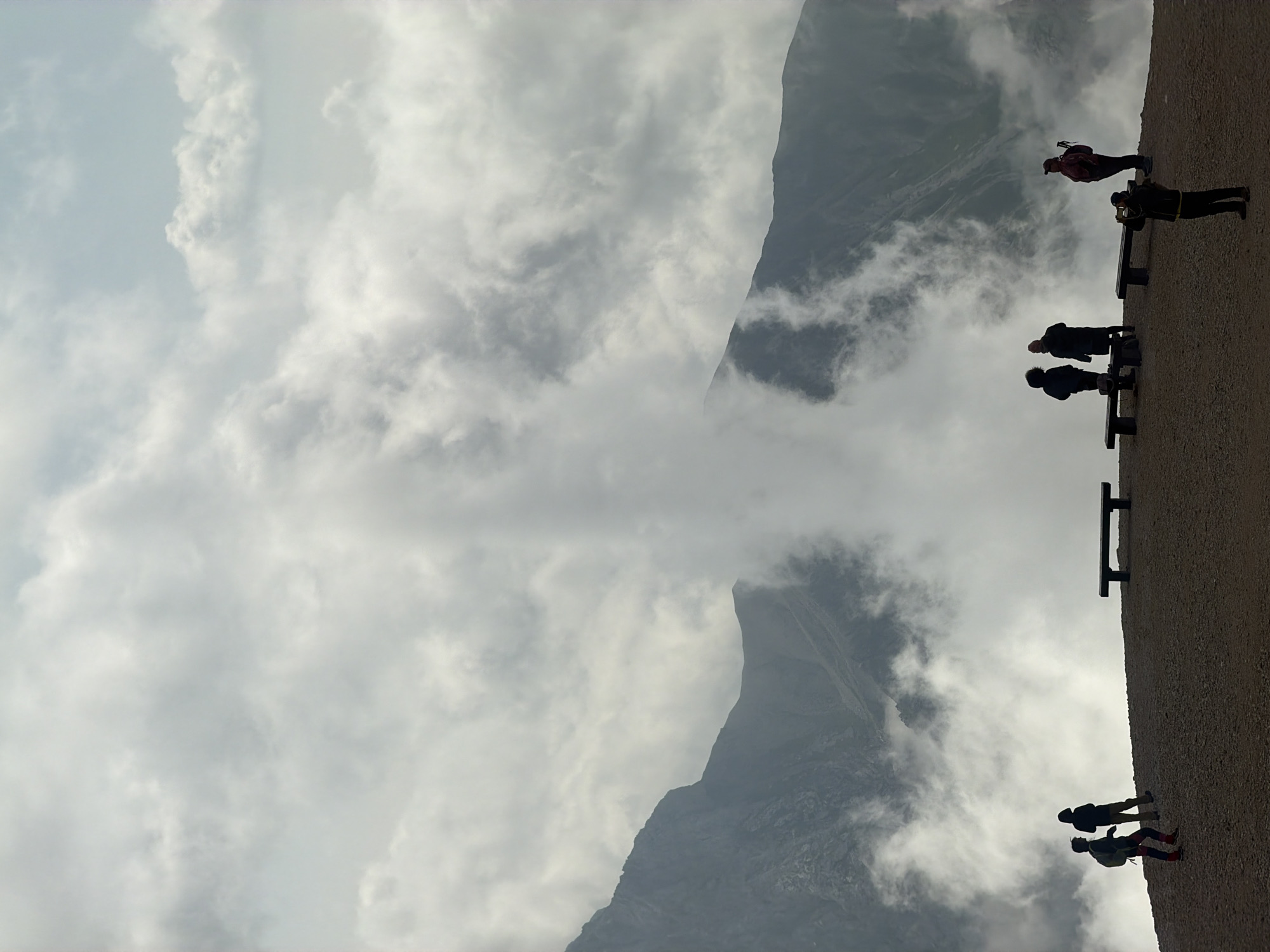 A panoramic photograph captures a dramatic mountain landscape shrouded in clouds. In the foreground, a dusty, sloped terrain is dotted with several silhouetted figures. These individuals, some standing and others seated on dark, minimalist benches, are observing the misty expanse before them. One person is actively taking a photograph. The mountains themselves are rugged and steep, with the cloud cover creating an ethereal, almost otherworldly atmosphere. A prominent column of mist rises vertically from the mountain face, adding to the scene's mystique. The overall mood is one of awe and contemplation, with the raw beauty of nature on full display. The image is located in the municipality of Wildermieming, Austria.