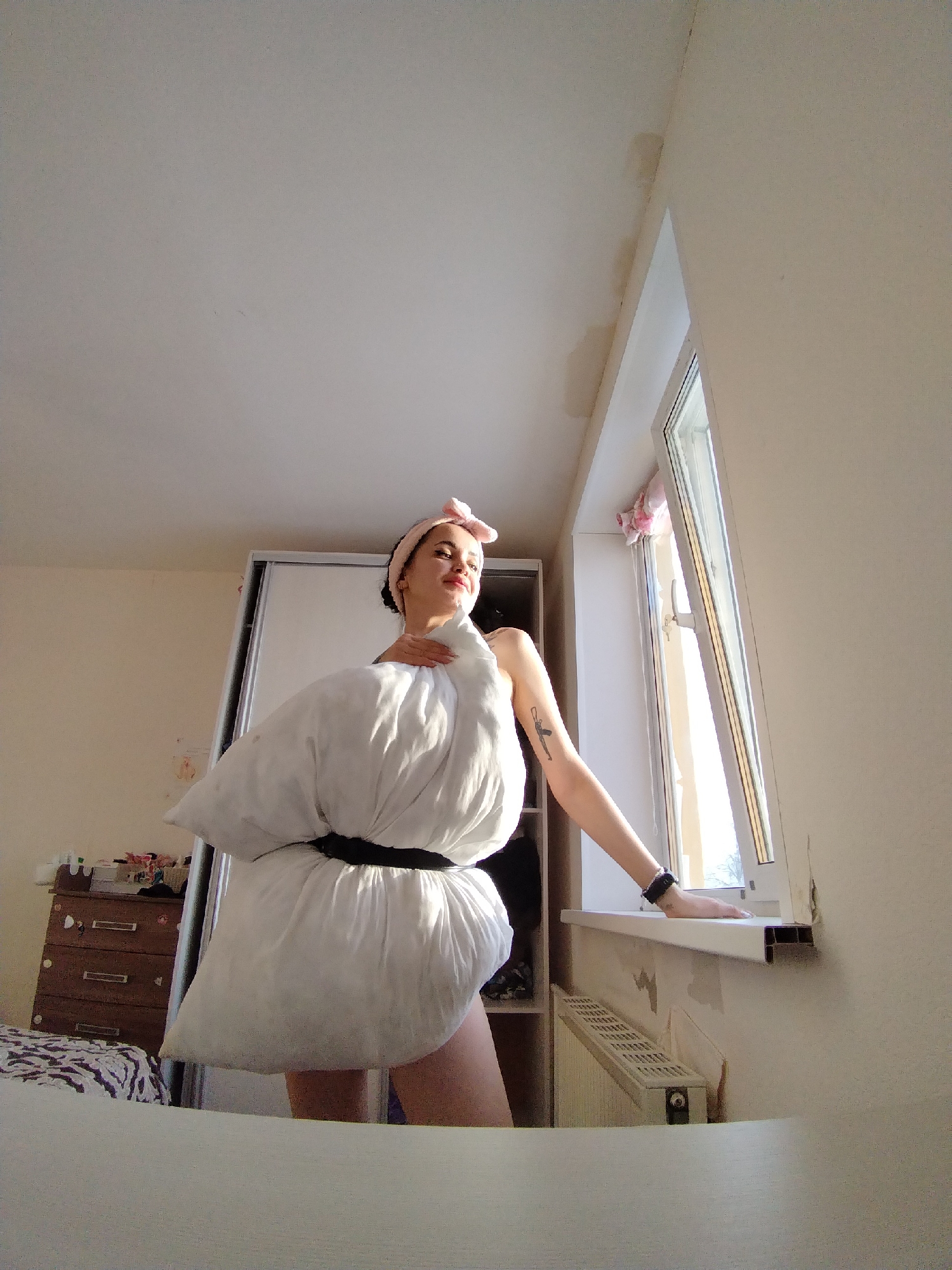 A young woman stands indoors wearing a pillow secured around her waist with a black belt, posing for a photograph. She has a pink headband in her hair and a tattoo on her left arm. Her right hand rests on the windowsill of an open window.
The room features a white wardrobe, a wooden dresser, and a radiator beneath the window. The wall shows signs of peeling paint. The image is taken from a low angle, capturing the ceiling. The location might be Noviy, Russia, based on the request.