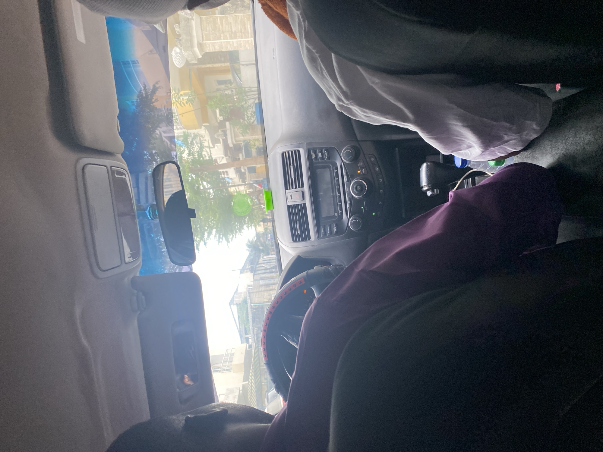 The image displays the interior of a car, viewed from a low perspective looking towards the front windshield. A person is partially visible in the bottom left foreground, wearing a dark top. An arm and torso, clothed in a purple long-sleeved garment, are also visible on the right side of the frame, near the steering wheel and center console, suggesting this is the driver. In the upper right corner, the legs and lap of another person are visible, covered by a white sheet or blanket, indicating a passenger.
The car's dashboard, steering wheel, and center console are prominent. The black steering wheel is fitted with a red and black cover, and an orange indicator light glows on the dashboard behind it. The car's audio system, featuring controls labeled "AM/FM," "CD," and "AUX," along with climate controls and various knobs, is visible on the center stack. A gear shifter is present, and two plastic bottles (one with a green cap, one with a blue cap) are nestled in the console area beside a white fabric item. A rearview mirror is in view, and sun visors are seen above the windshield, with one lowered.
Through the windshield, a bright, sunny daytime scene unfolds. Buildings with light-colored and yellow facades, interspersed with green trees, are visible, suggesting an urban or residential environment. A circular green sticker is affixed to the windshield. The car appears to be stationary on a clear day.