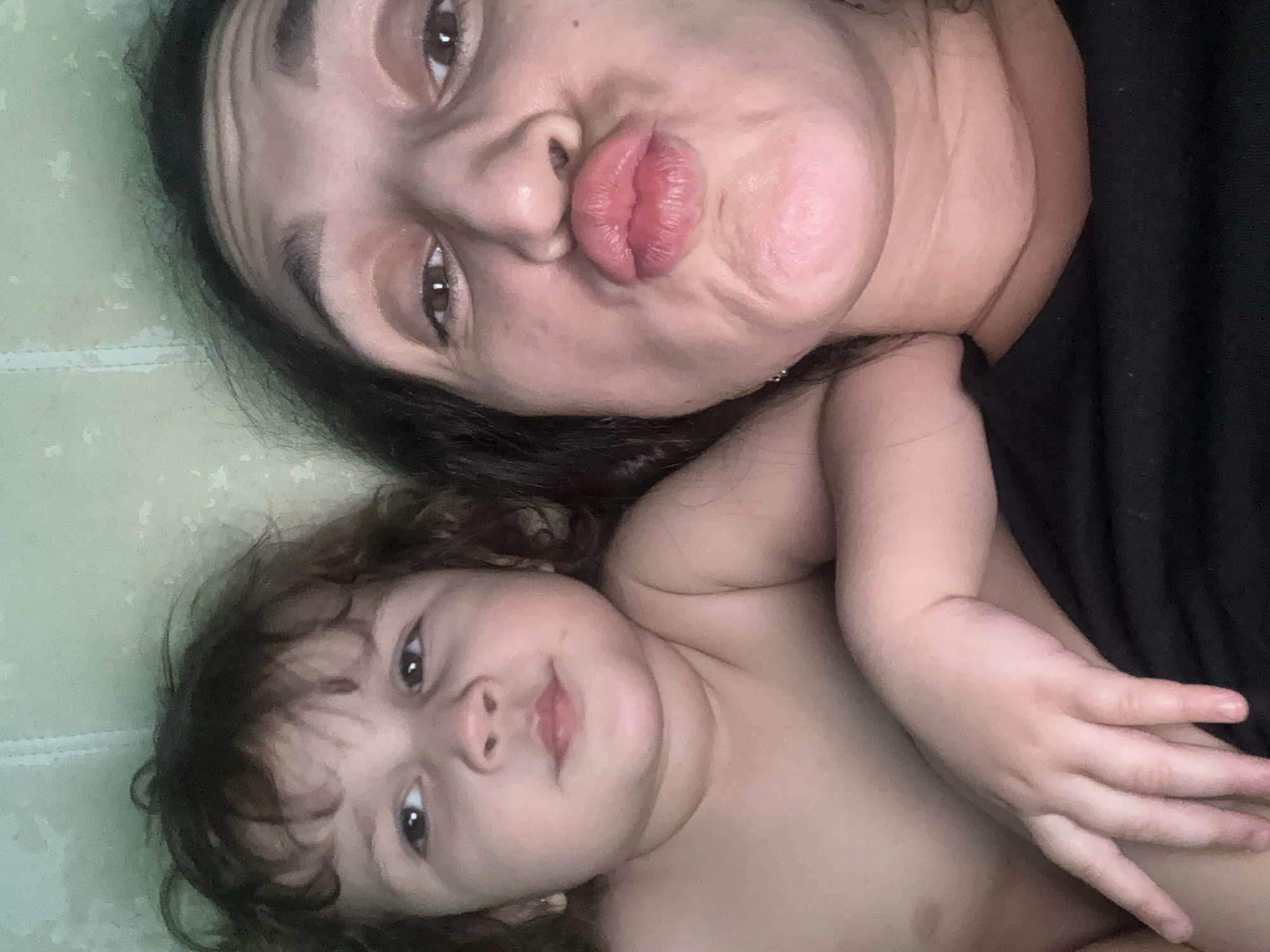 A close-up, rotated image captures an adult and a young child lying side-by-side on a light green surface, likely a bed or blanket, in what is contextually identified as Tobolsk, Russia. The image is rotated 90 degrees clockwise, positioning the subjects' heads towards the upper left and their bodies extending towards the lower right.
The adult, occupying the upper portion of the frame, has dark hair and dark eyes. She is making a playful "kiss" face with prominent, puckered lips. Her skin tone is medium, and she is partially dressed in what appears to be a dark-colored garment visible on the right side of the frame.
Adjacent to and below the adult is a young child with curly dark hair and dark eyes. The child looks directly at the camera with a soft, curious, or neutral expression. The child is bare-chested, revealing fair skin and a visible arm and hand.
The background is a consistent light green with faint, irregular lighter speckles or patterns, suggesting a textured fabric. A subtle horizontal line, possibly a fold or seam in the fabric, runs across the background near the adult's head. The overall mood of the image is intimate, affectionate, and playful. No text is visible in the image, and details such as time of day or weather cannot be determined.
