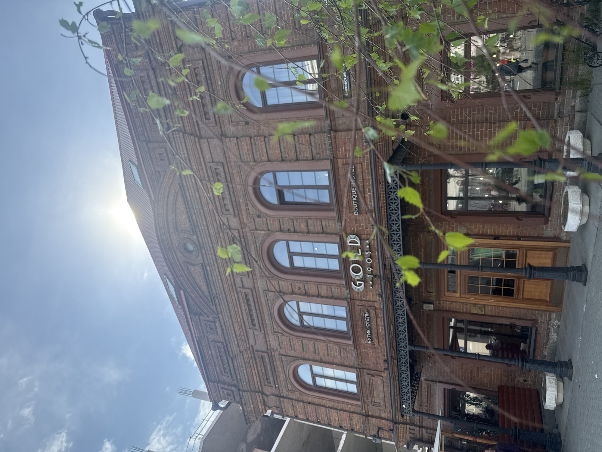 The image shows the exterior of the boutique hotel "Gold 1905" in Yekaterinburg, Russia. The building is made of red brick with arched windows. There is a sign on the building that says "GOLD 1905" and "БУТИК-ОТЕЛЬ" (Boutique Hotel) in Russian. There are decorative metal supports near the entrance. Tree branches and leaves are partially obscuring the view. The sky is blue with some clouds. The sun is shining brightly, causing a glare on the upper part of the building. There appears to be construction occurring to the left of the building. A pedestrian walkway is visible in the foreground.