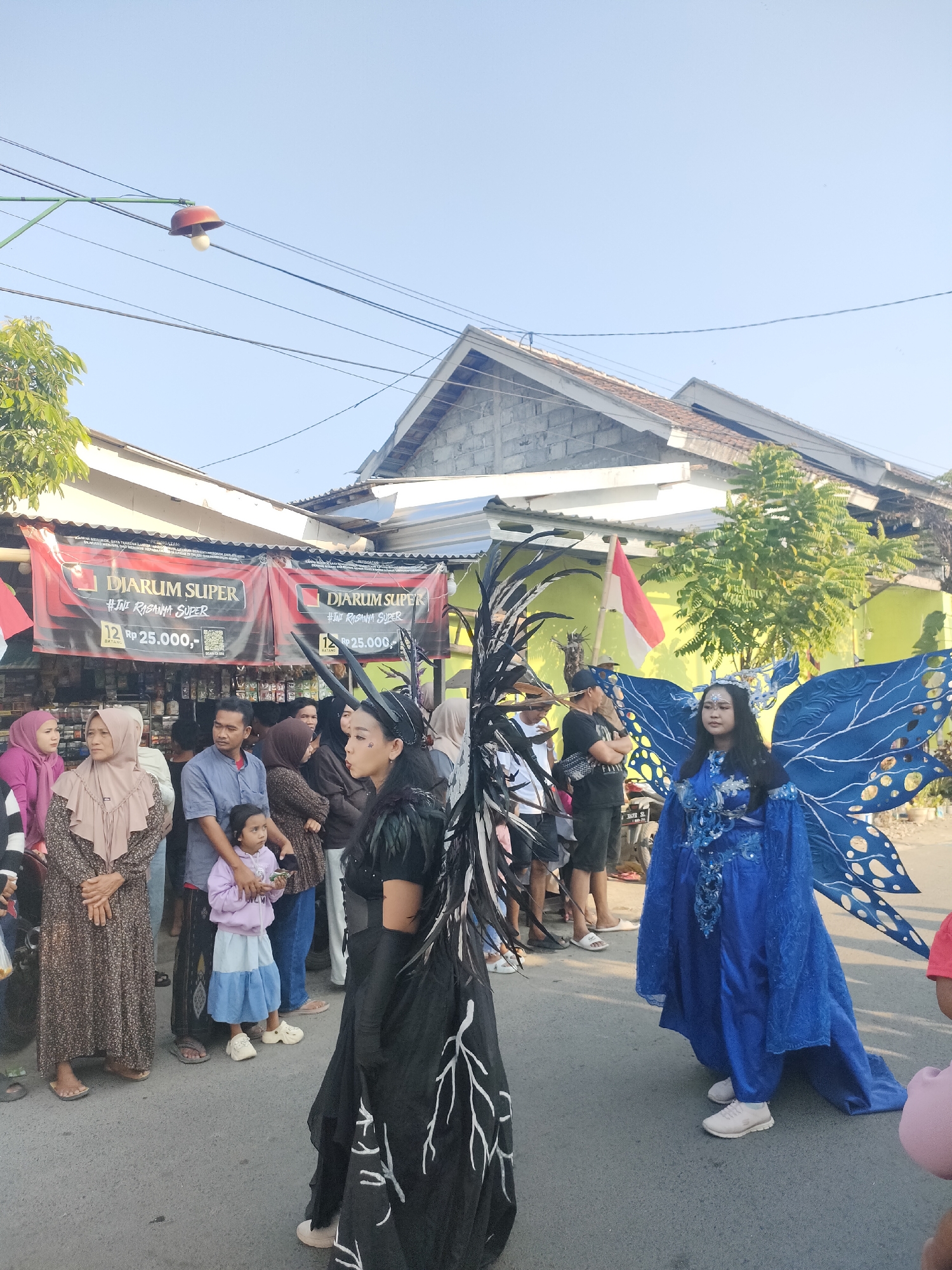 In Tongaswetan, Indonesia, a street scene unfolds under a clear sky, likely during a daytime event. The foreground is dominated by two individuals in elaborate costumes. To the left, a person wears a dark, feathered costume with prominent black wing-like structures and a horned headpiece. To the right, another person is dressed in a vibrant blue gown adorned with sparkling embellishments, complemented by large, intricately designed blue wings that extend outwards.
Behind them, a crowd of onlookers lines the asphalt road. Many are dressed in casual attire, with some women wearing hijabs. Several people are standing, while others appear to be watching the costumed individuals. A banner for "Diarum Super" is visible behind the crowd, featuring text and prices. Red and white Indonesian flags are also displayed. The background shows buildings with tiled roofs and some greenery, suggesting a residential or commercial area. The overall atmosphere appears festive and public.