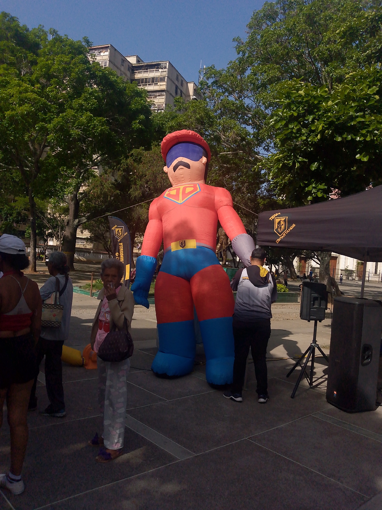 The image depicts a public space in Caracas, Venezuela. A large, inflatable superhero figure, predominantly red and blue, stands prominently in the foreground. Several people are visible around the figure: an elderly woman is smoking and is partially obscured by the figure; a person stands near a speaker and tent marked "Team Espartanos"; two individuals with shopping bags are partially obscured. The background includes trees, a building, and vendor tents. The scene appears to be during the daytime, with clear skies. The ground is paved with dark tiles. The lighting suggests a sunny day.