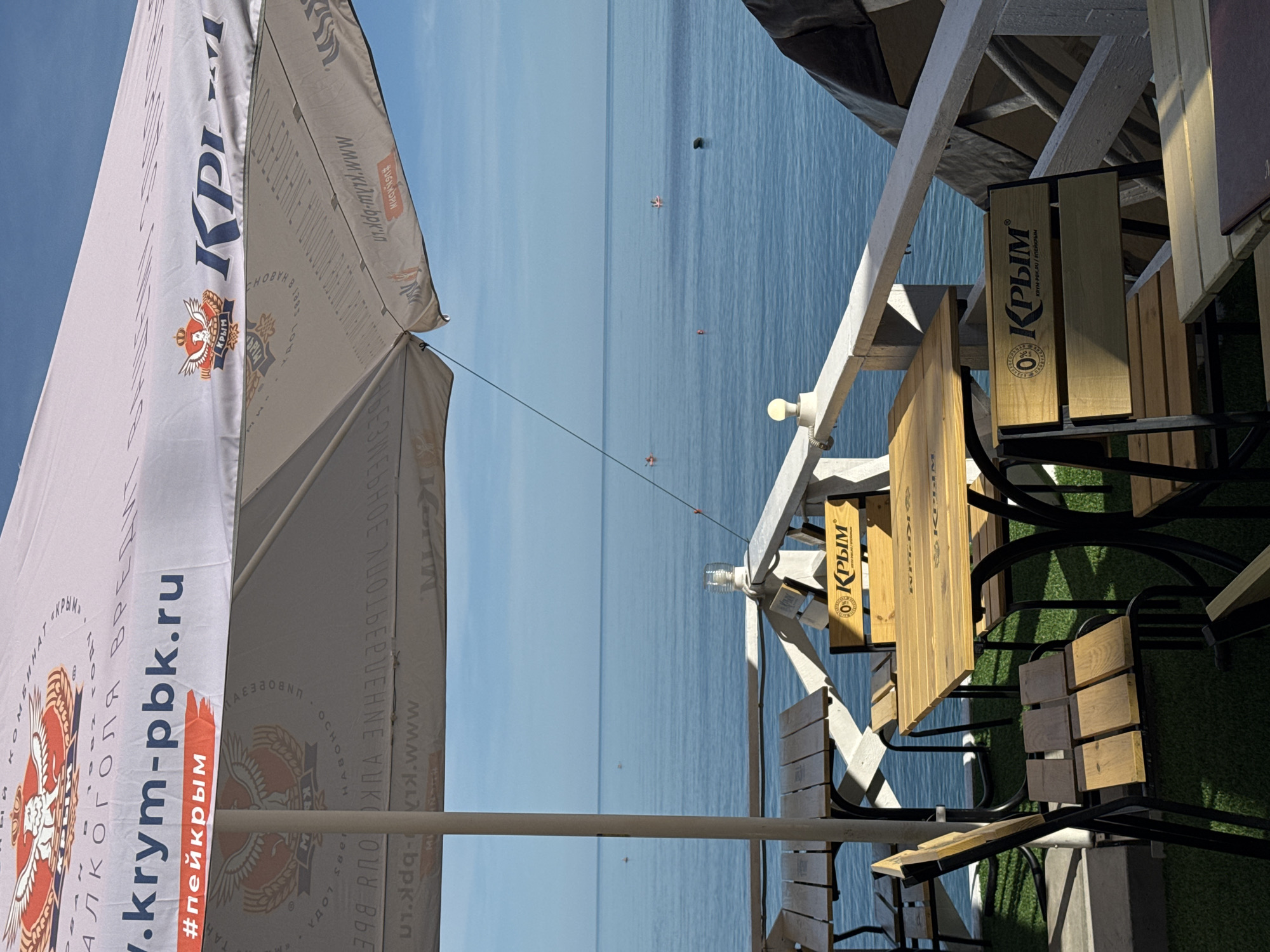 The image shows an outdoor seating area with wooden tables and chairs on a terrace overlooking the sea. The sky is clear and blue, and the water is calm. An umbrella, presumably for shade, is set up over the tables. The umbrella has logos and text printed on it, including "krym-pbk.ru" and "#пейкрым." The seating area is situated on what appears to be a green carpeted surface, suggesting it's part of a cafe or restaurant. In the distance, there are a few small boats on the water. The overall scene depicts a tranquil seaside location.