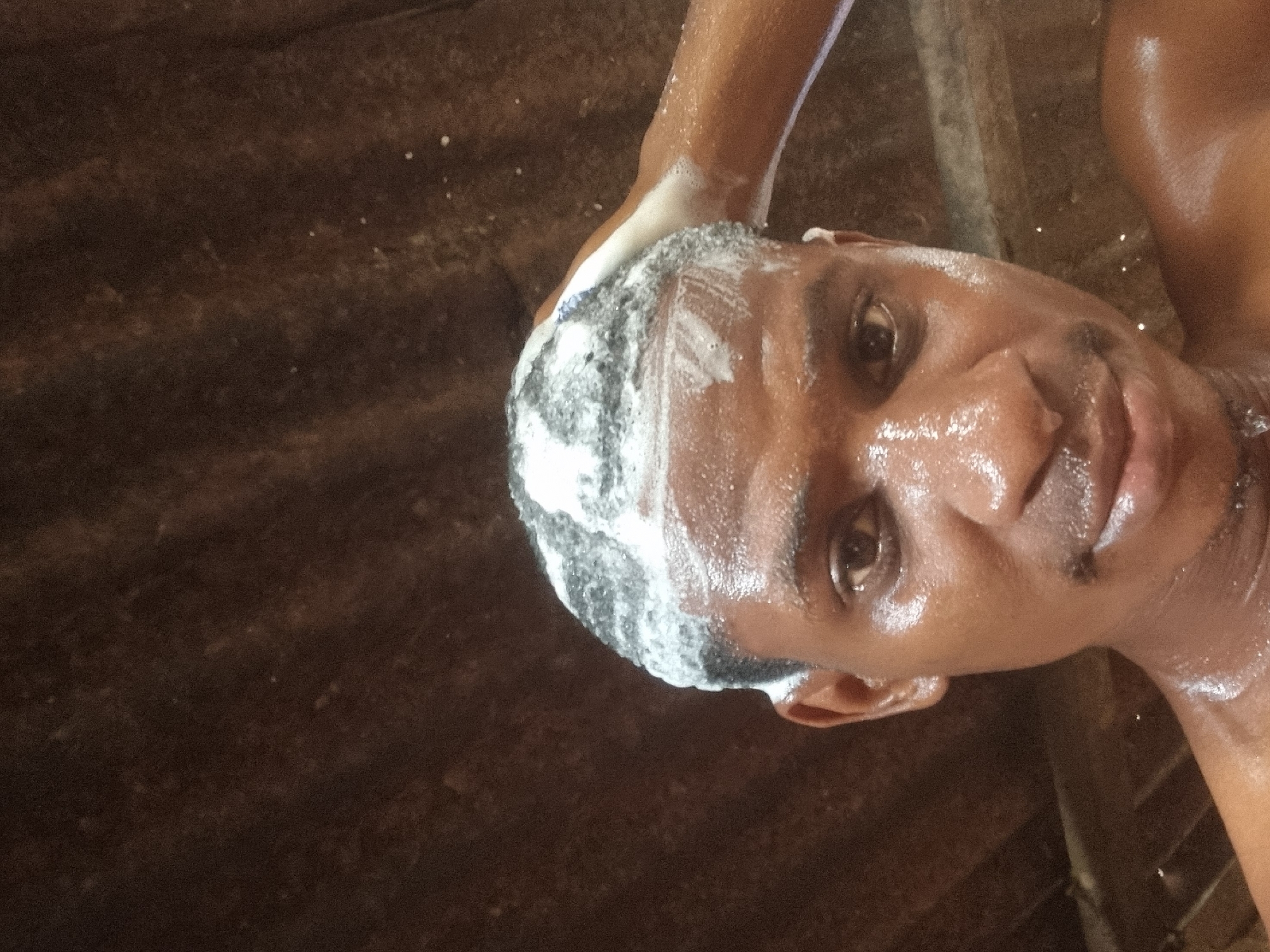 A dark-skinned man is washing his hair with soap or shampoo in what appears to be an outdoor or semi-outdoor setting. The location in Onitsha, Nigeria cannot be confirmed, though the setting may resemble a typical bathing area in a less urbanized part of the city. The man has short, dark hair, and his skin is wet and reflective. He is looking directly at the camera with a neutral expression. The background is dark and indistinct, possibly a wooden structure or wall. A portion of an arm is visible, presumably belonging to the man. The presence of scattered white specks may be residues of the washing product.