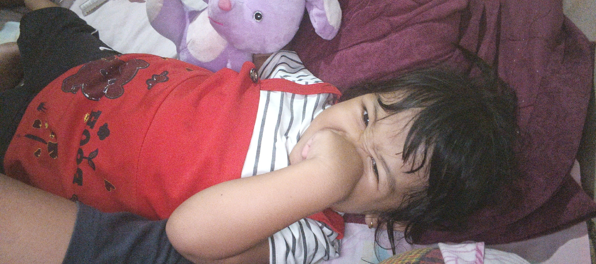 A child is lying on their back on a bed with a purple blanket. The child is wearing a red shirt with black and white stripes on the sleeves and collar. The child has black hair and is looking at the camera with their right hand in their mouth. A purple stuffed animal is lying on the bed behind the child. The image appears to have been taken in a bedroom in Cinta Dame, Indonesia.