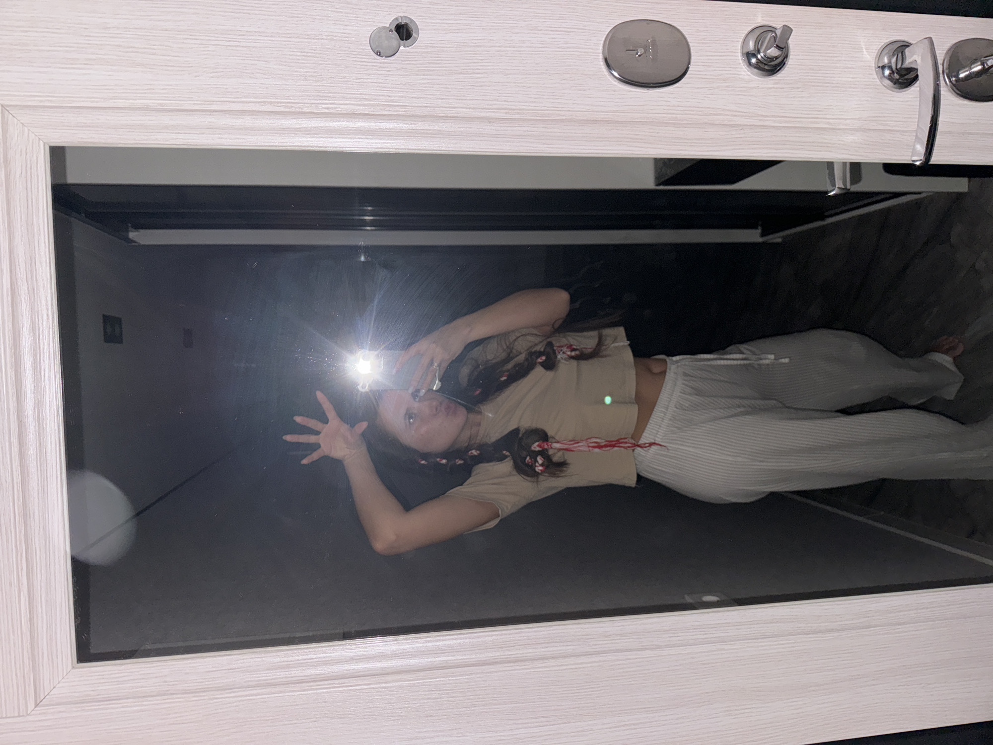A reflection in a framed mirror shows a young woman taking a selfie. She is wearing a cropped beige top and white striped pants. Her hair is styled with braids and red accents. The background reflected in the mirror appears to be a dimly lit room. The mirror frame is light-colored with a wood grain pattern, and various metal hardware elements are attached to the top of the frame. The location is Unknown, Unknown.