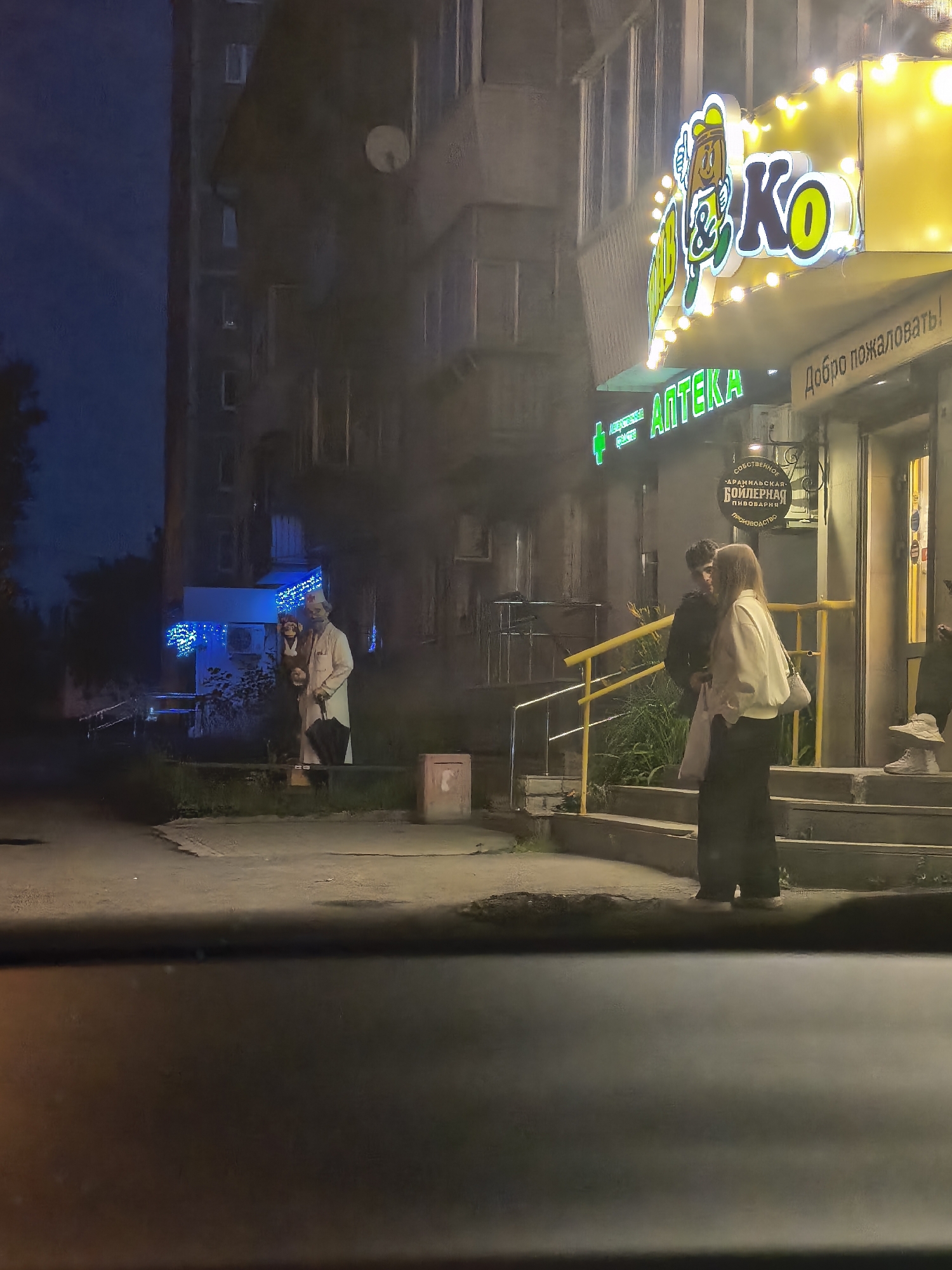 The image depicts an outdoor nighttime scene in front of an establishment with a bright yellow sign that reads "КО & АПТЕКА" in green letters. The sign features a cartoon character. Below the main sign, there's a smaller sign that says "Добро пожаловать!". To the right of the entrance, there are steps leading up to the building. A couple is standing on the steps, facing each other, with the male wearing a black jacket and the female wearing a white jacket.
On the left side of the image, further from the building, are two figures standing near some blue lights. One figure appears to be wearing a white coat and holding an umbrella, while the other figure is smaller and seems to be wearing a dark outfit.
The environment suggests a city street, with a building in the background and a road or pavement in the foreground. The lighting is primarily from the illuminated signs and building lights, creating a somewhat hazy atmosphere.