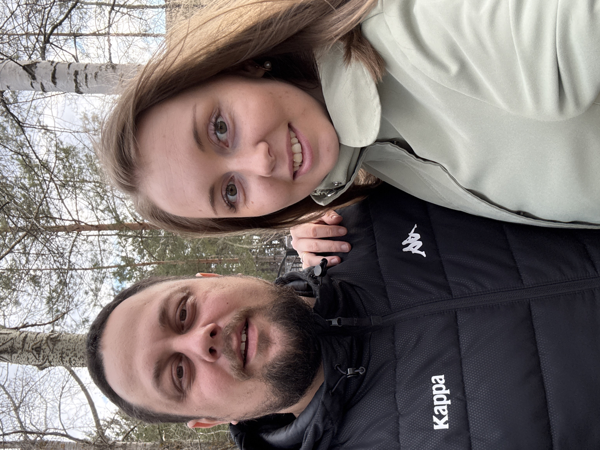 This rotated image captures a close-up selfie of a man and a woman outdoors in a wooded setting. The image is oriented with the subjects' heads to the right and their bodies extending downwards.
The woman, positioned above the man (or to his right if the image were correctly oriented), has long, light brown hair and is smiling towards the camera. She wears a light green jacket and has an earring visible on her right ear. Her right hand rests gently on the man's shoulder/jacket, revealing a silver ring on her ring finger.
The man, beneath the woman (or to her left), has a full beard, short dark hair, and is also smiling. He wears a black padded jacket. On his jacket, the white Kappa logo, depicting two figures leaning against each other, is clearly visible, as is the word "Kappa" printed vertically in white.
The background shows a sparse forest or woodland, dominated by bare trees, including distinctive birch trees with white bark. The sky appears bright but partly cloudy, suggesting cool weather typical of late autumn, winter, or early spring. Both individuals appear happy and content, engaged in taking a shared photograph.