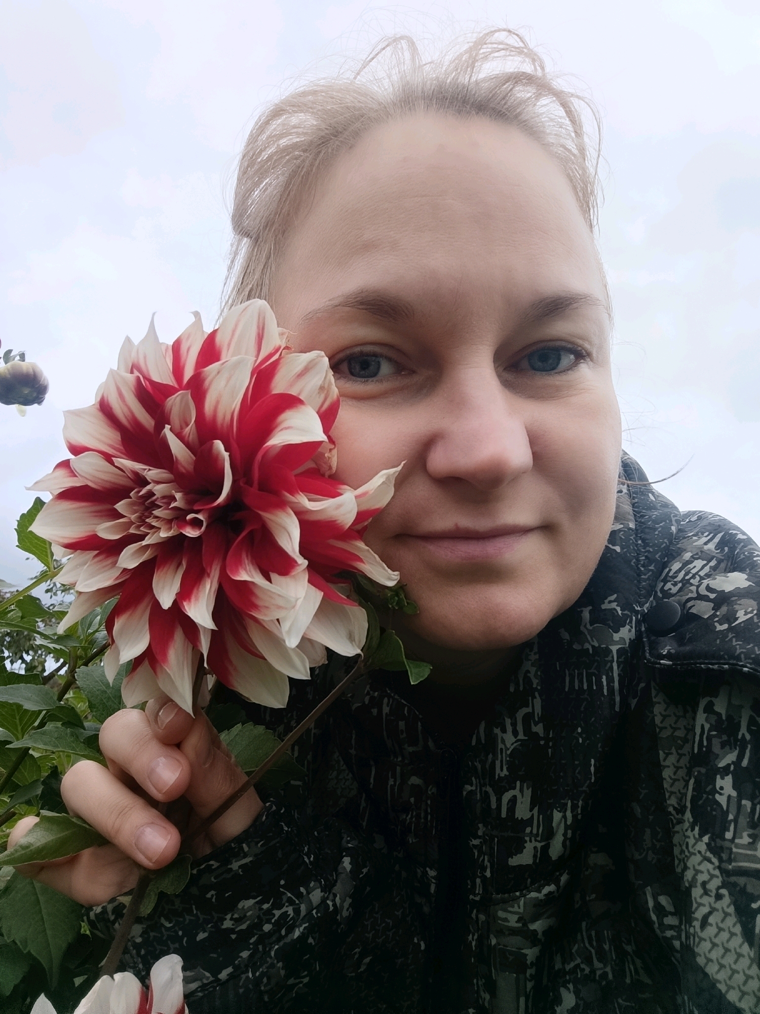 A medium-shot, selfie-style photograph shows a fair-skinned woman with blonde hair pulled back, smiling slightly as she holds a large, red and white dahlia flower in front of her face. Her right eye is visible behind the flower, and her left eye is slightly obscured. She is wearing a dark, patterned jacket. The background consists of green foliage and a cloudy sky. The image appears to have been taken outdoors, likely in a garden setting, potentially in the city of Dubovaya, Russia, given the context provided.