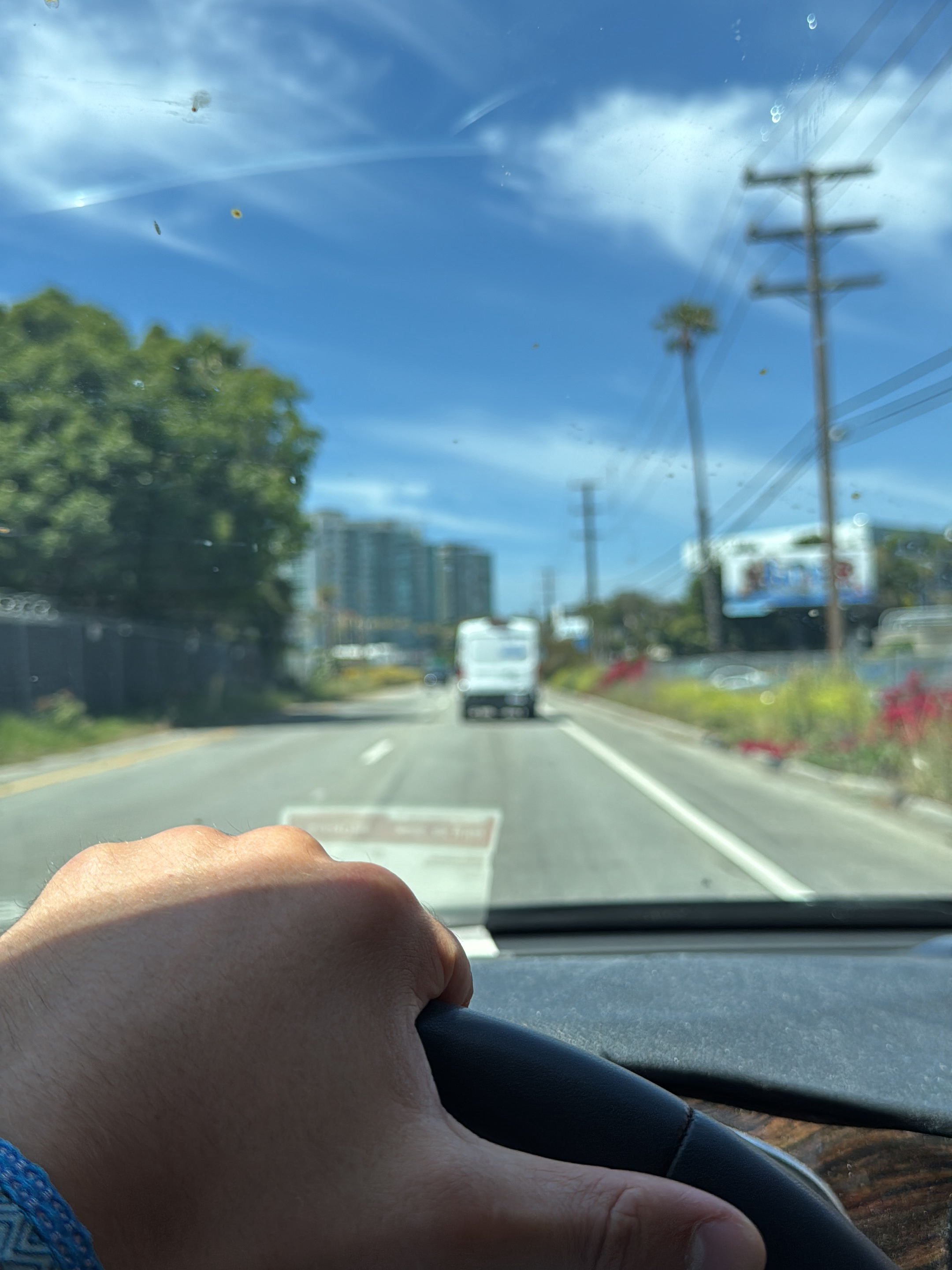 A photograph, taken from inside a vehicle, shows a driver's hand gripping the steering wheel. The vehicle is moving on a road with a white van visible in the distance. The scenery includes a combination of buildings, trees, and utility poles, indicative of an urban or suburban environment. The sky is clear with few scattered clouds, suggesting daylight hours and fair weather conditions. The location is Marina del Rey, United States.